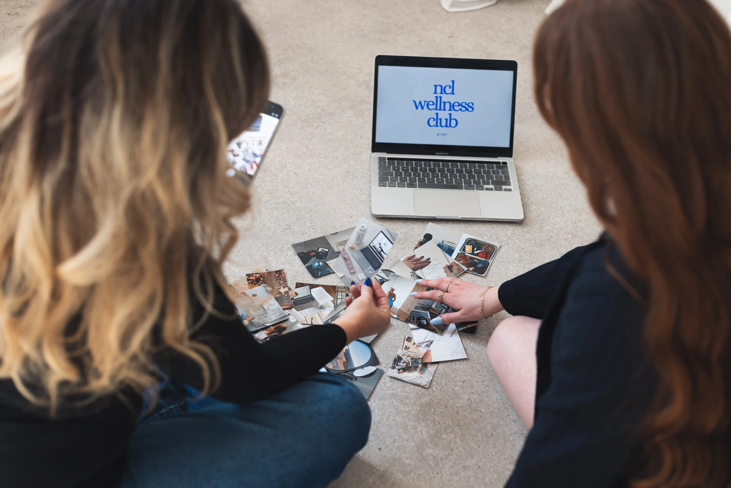 Two women with wavy hair sitting on the floor, looking at photographs, with a MacBook laptop displaying 'ncl wellness club' on the screen nearby.
