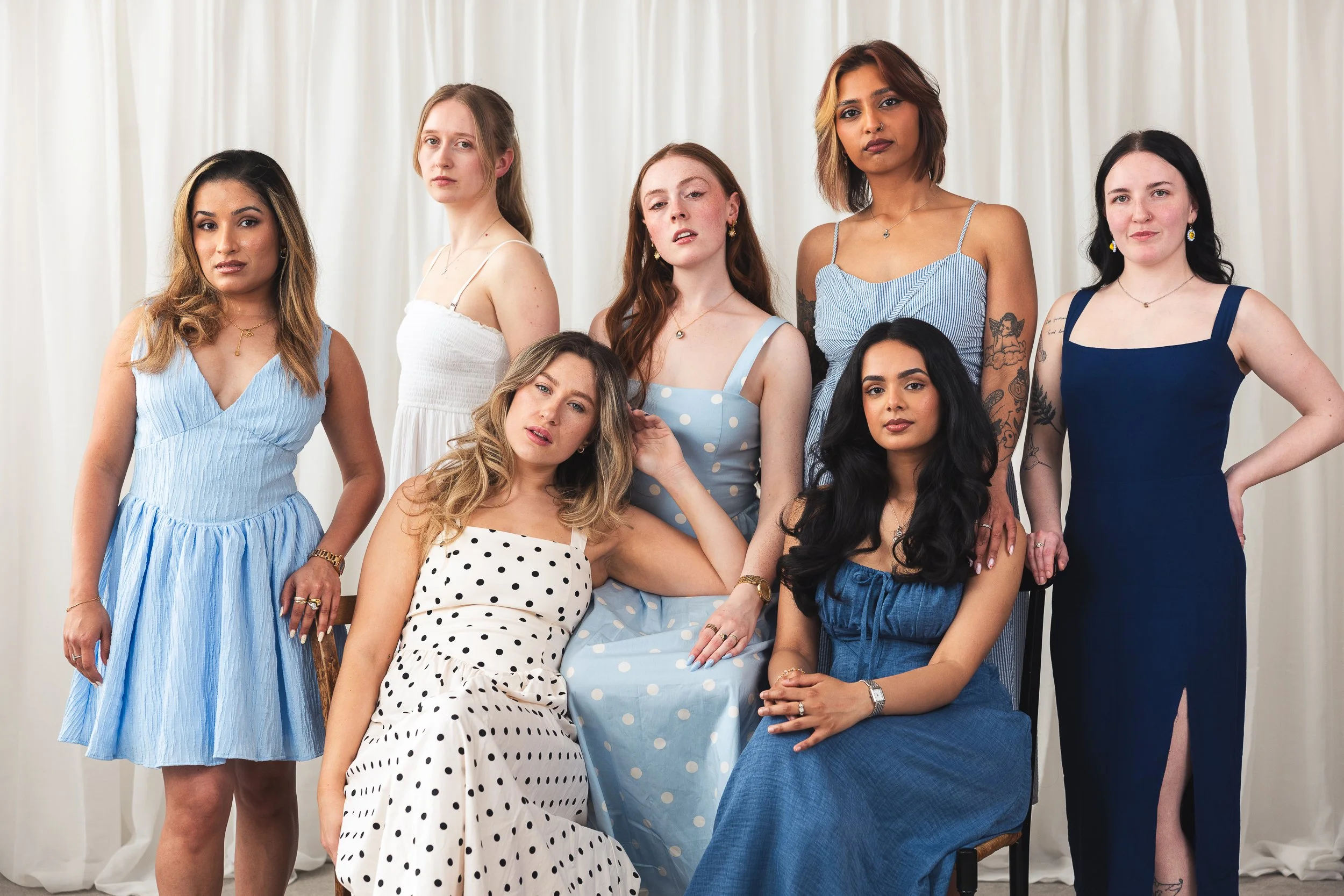 Group of seven women posing in front of a white curtain backdrop, wearing summer dresses in various colors and styles, with some seated and others standing.