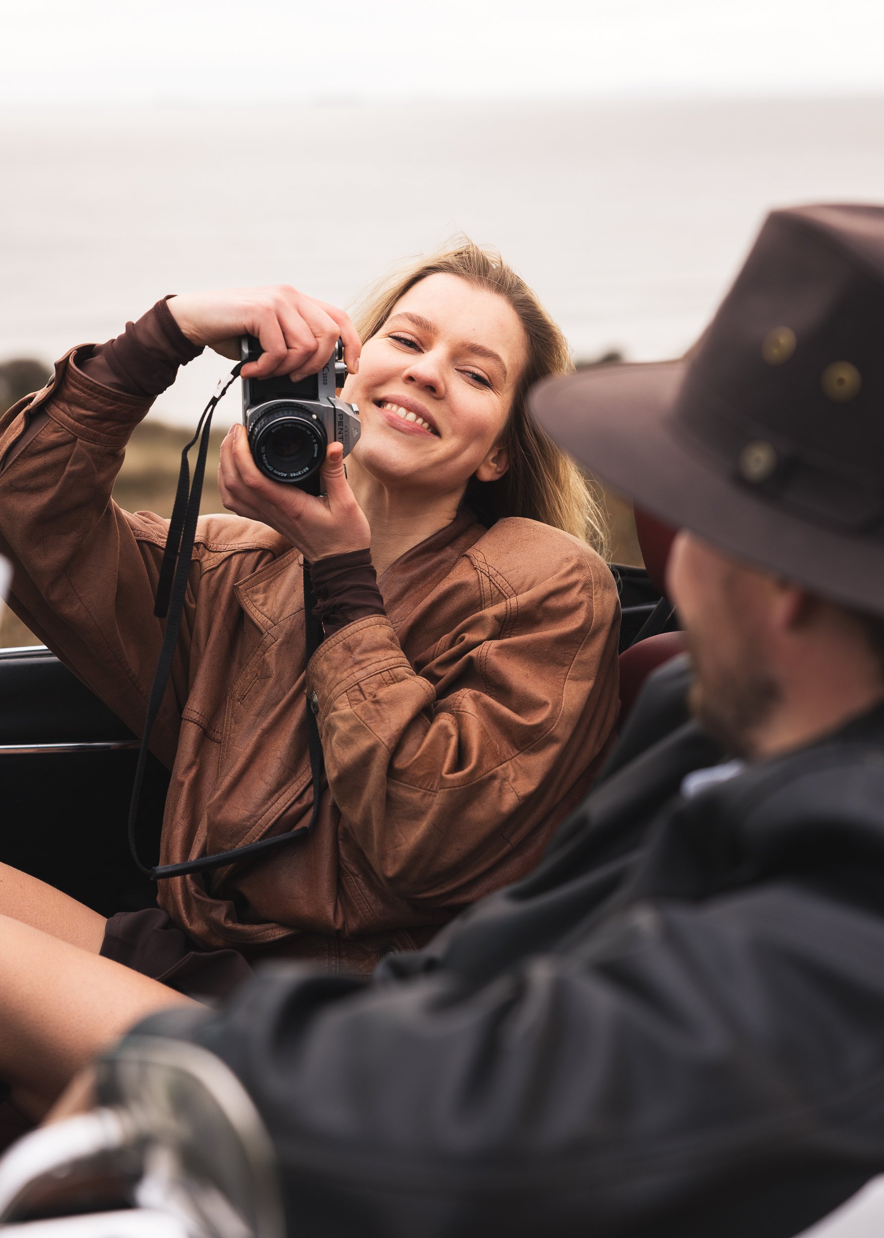 A woman smiling while taking a photo with a camera in a convertible car, with another person wearing a cowboy hat in the foreground.