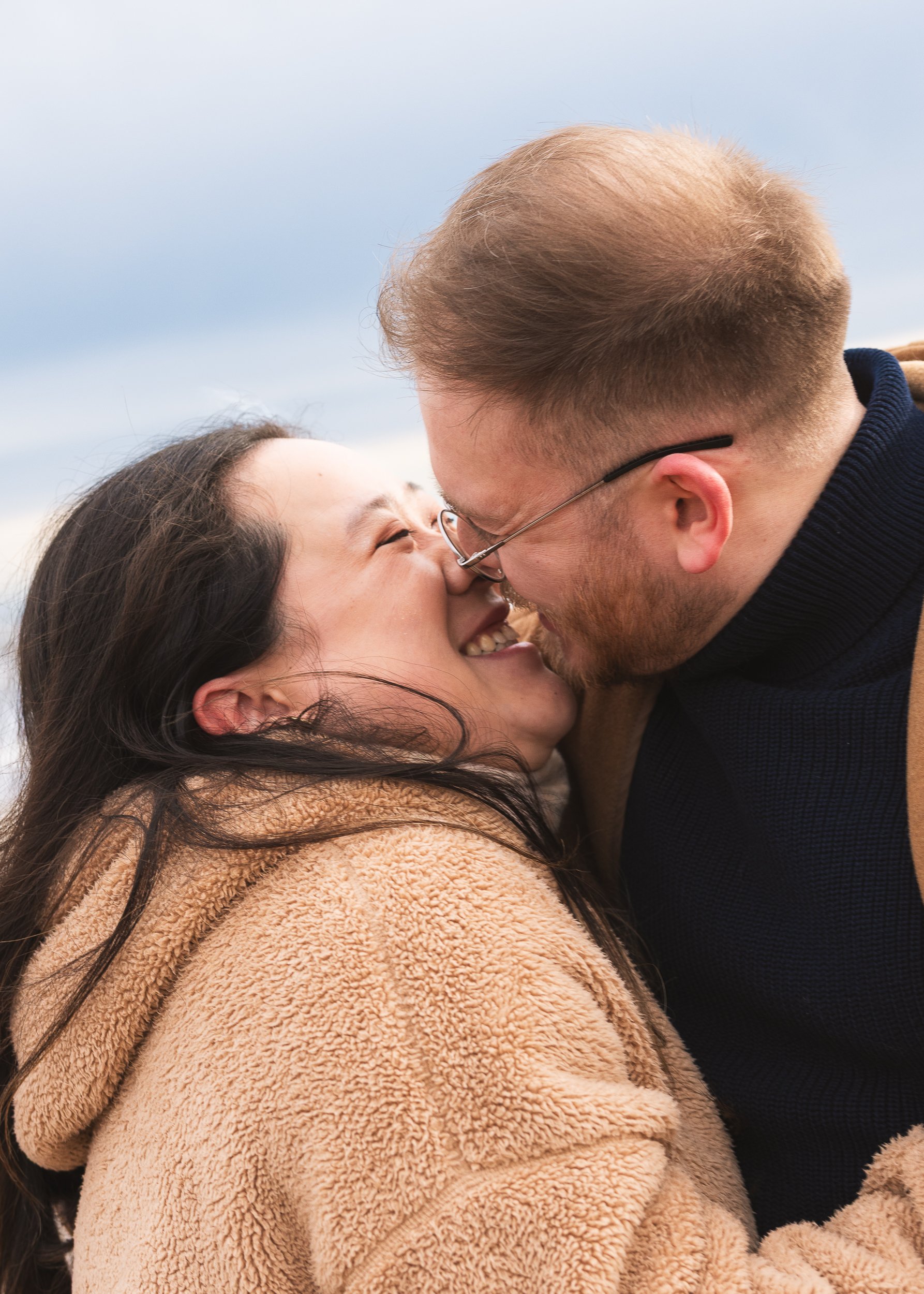 A couple kissing outdoors, woman wearing a brown fuzzy coat and the man wearing glasses and a dark jacket, against a cloudy sky.