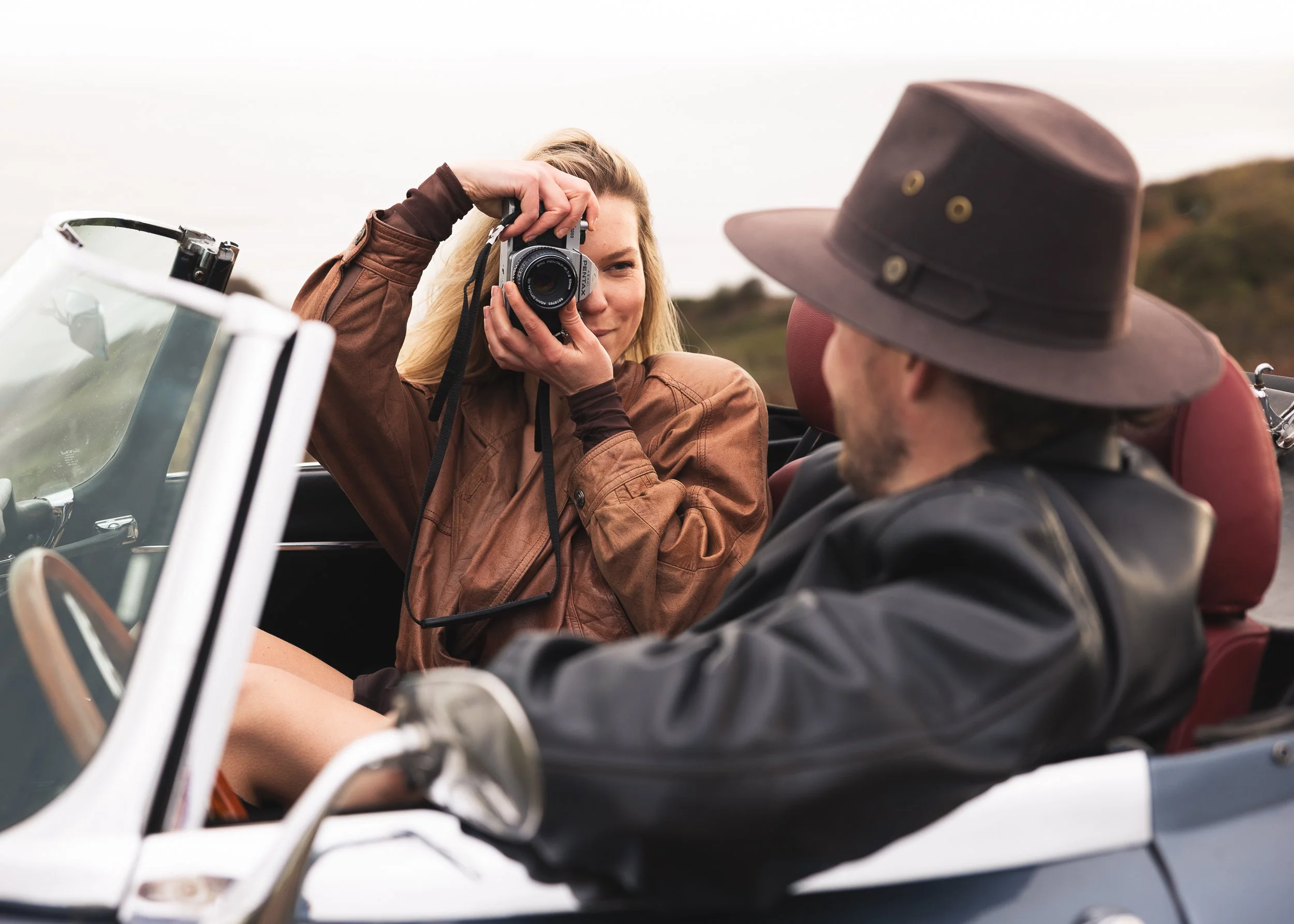A woman taking a photograph of a man in a vintage convertible car with a camera, with a hat and an outdoor landscape background.