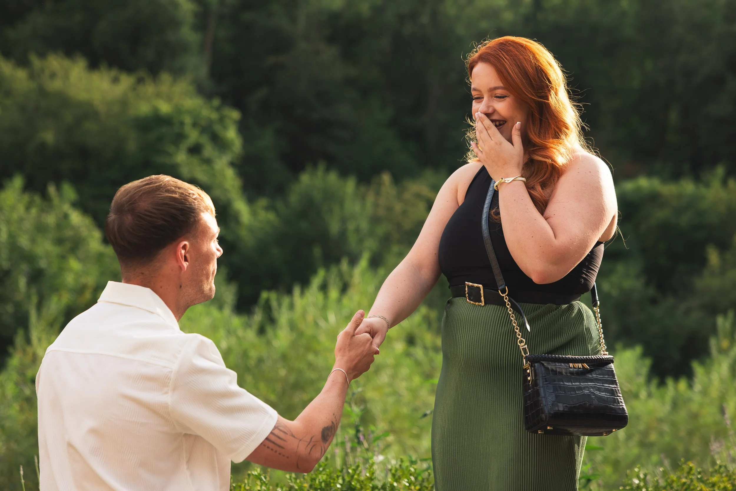 A man proposing marriage to a woman outdoors in a lush green setting. The woman has red hair and is smiling, covering her mouth in surprise. The man is kneeling, holding her hand.