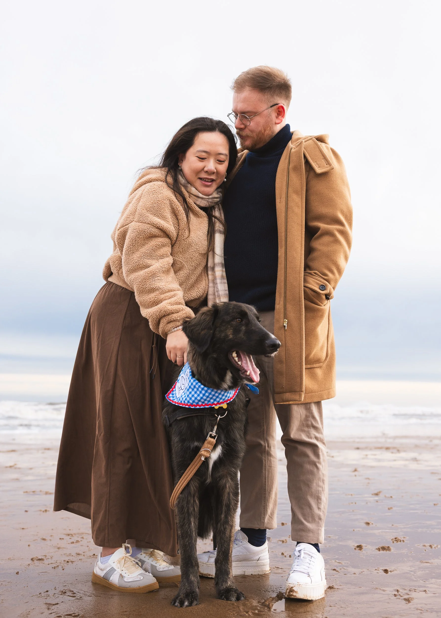 A couple with a dog on a beach, the woman is leaning on the man, and they are standing close together, with the dog in front of them.
