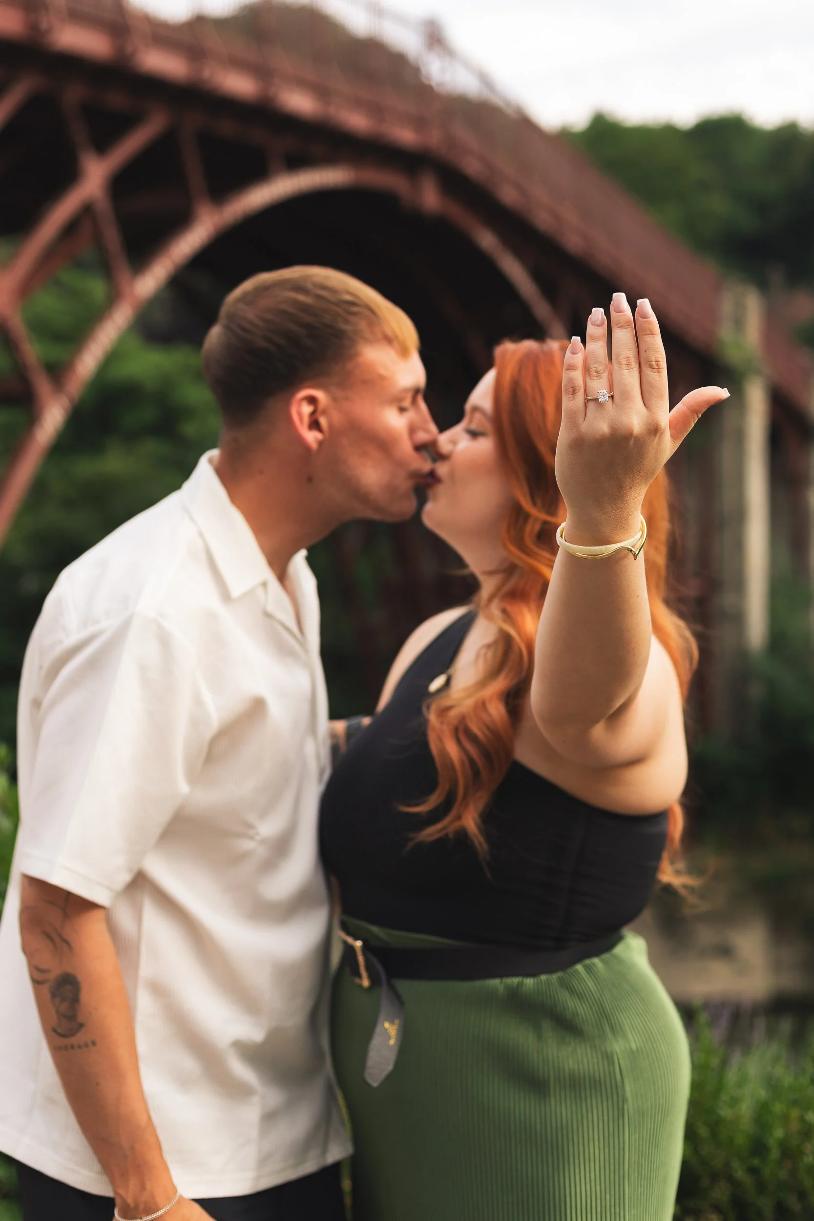 A couple kissing outdoors with a bridge in the background. The woman has red hair, is wearing a black top and a green skirt, and is displaying an engagement ring on her left hand.