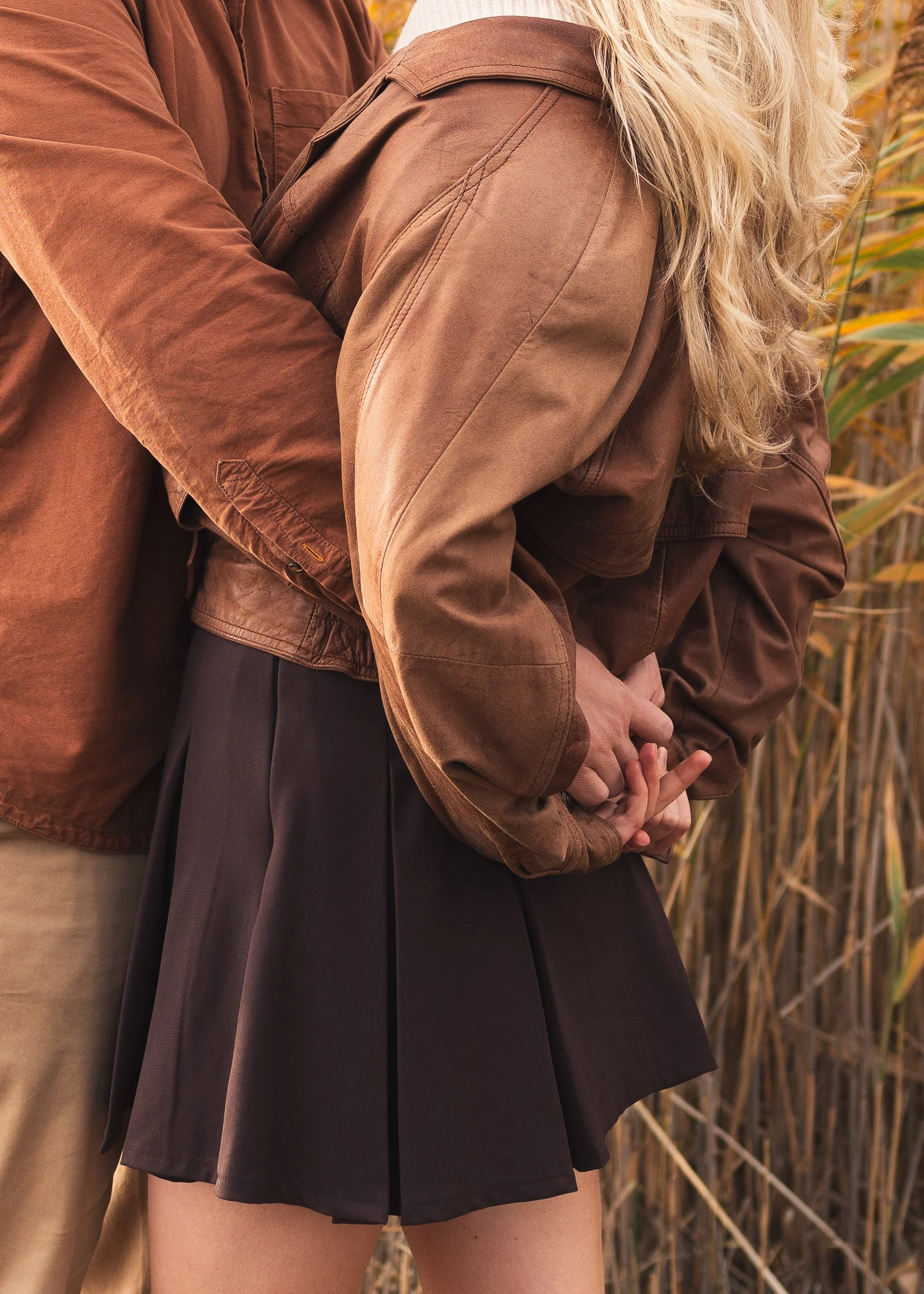 Close-up of two people holding hands, wearing brown jackets, standing outdoors near tall grass.