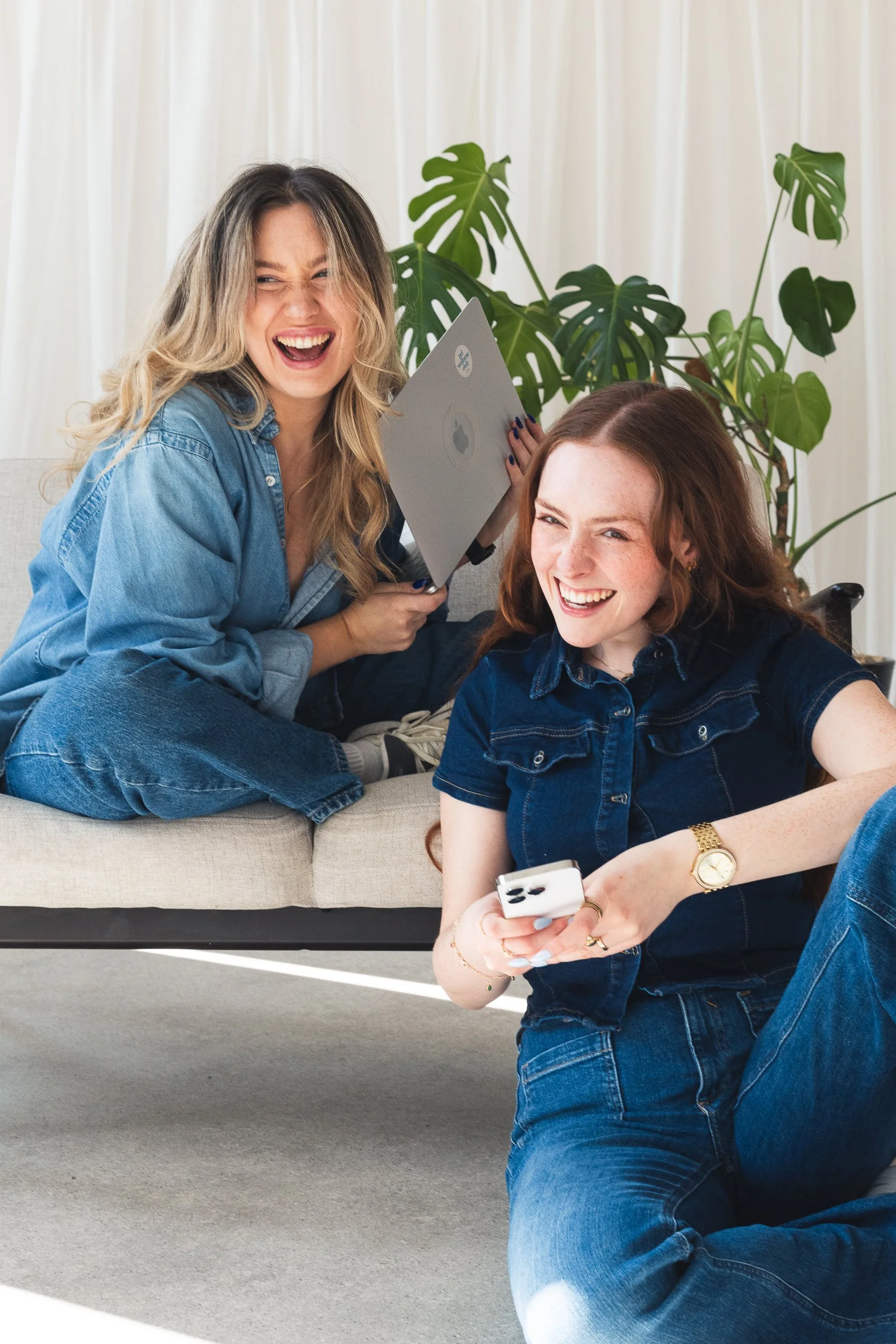 Two young women laughing and having fun, one is holding a tablet and the other is taking a selfie with a smartphone, with a large green plant and a white curtain in the background.