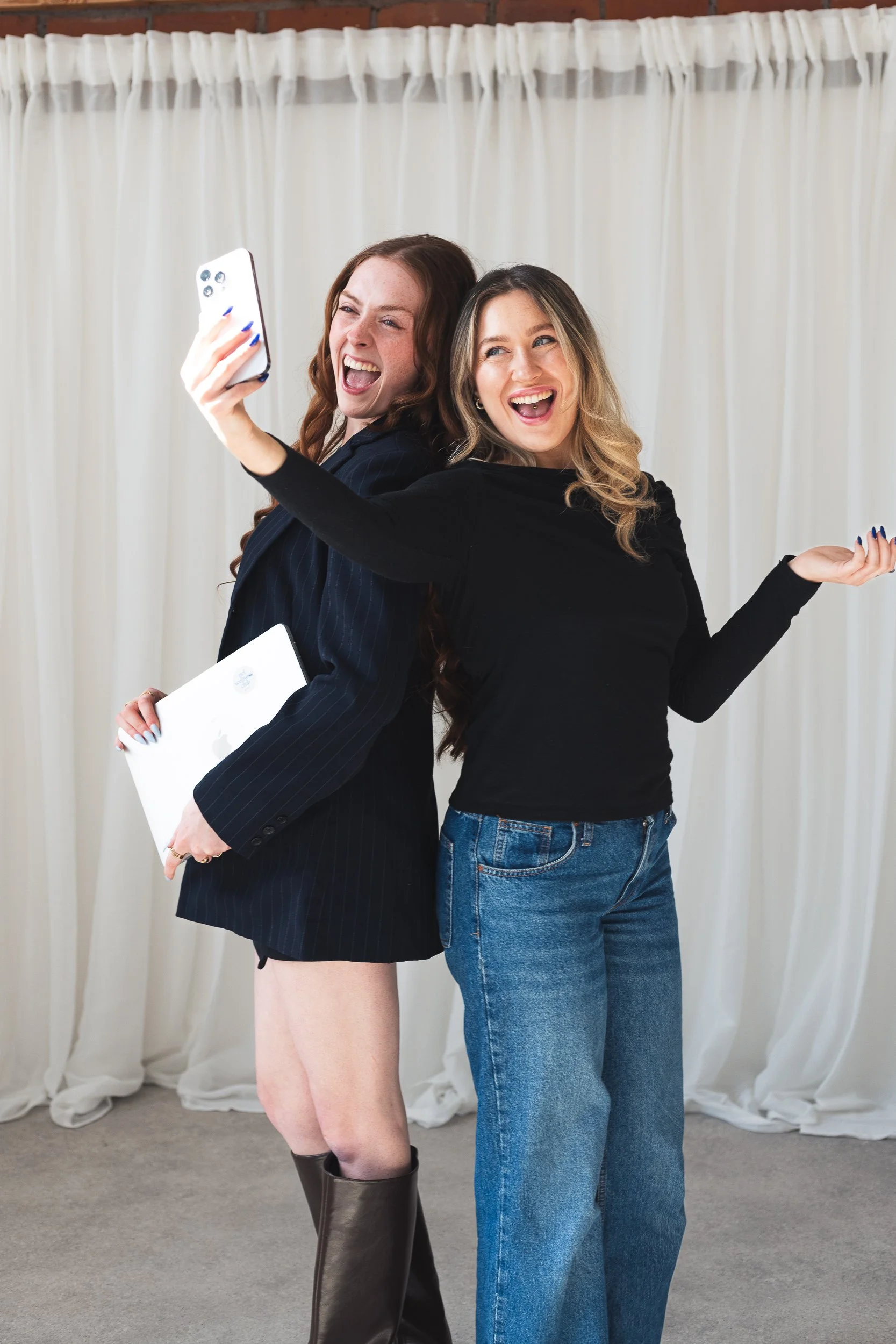 Two women taking a selfie together in front of a cream-colored curtain. One woman has long red hair, is wearing a black blazer with shorts, and black boots. The other woman has shoulder-length blonde hair, is wearing a black long-sleeve top and blue 