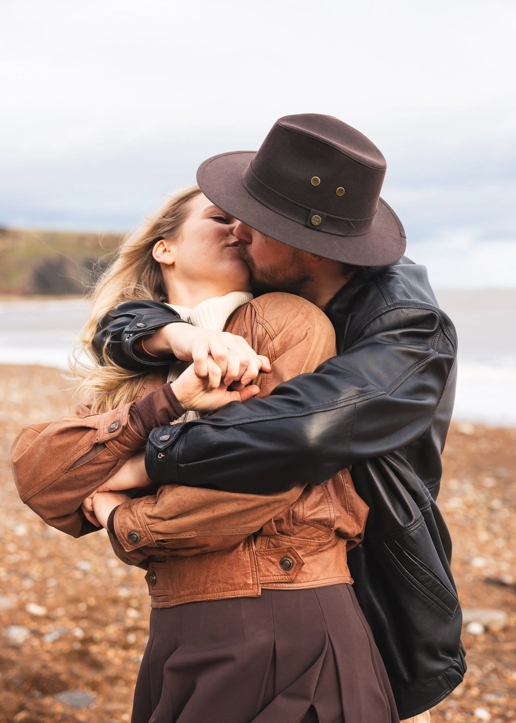 A man and woman hugging and kissing outdoors on a cloudy day, with a beach and cliffs in the background.