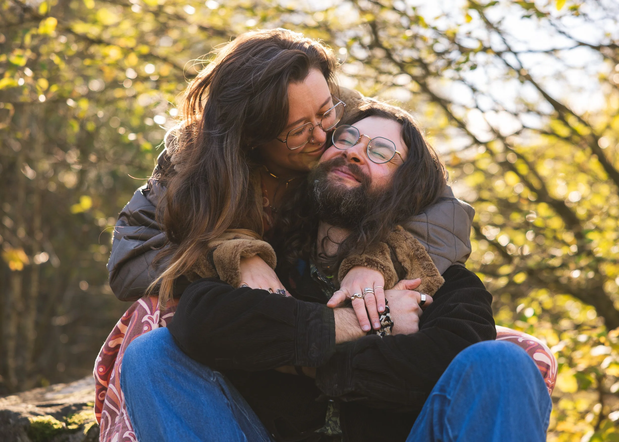 A couple hugging outdoors in a park during autumn, smiling with eyes closed, surrounded by trees with yellow leaves.