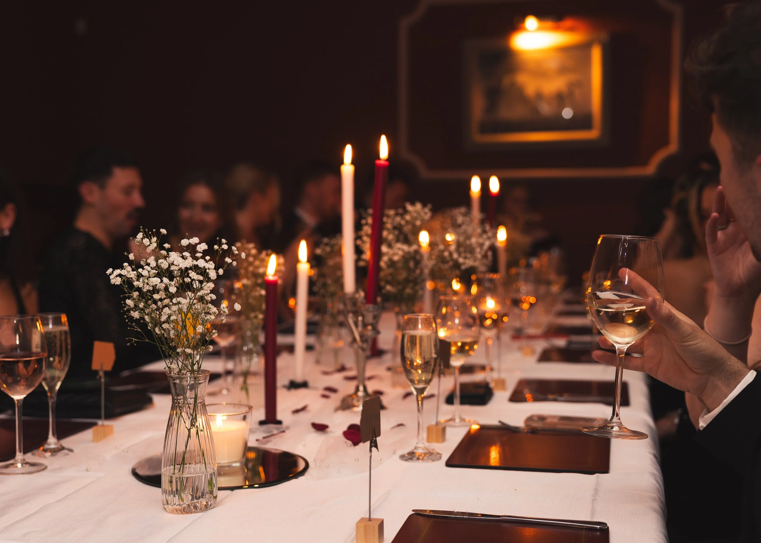 A formal dinner event with guests seated at a long table decorated with candles, flowers, and wine glasses in a dimly lit room.