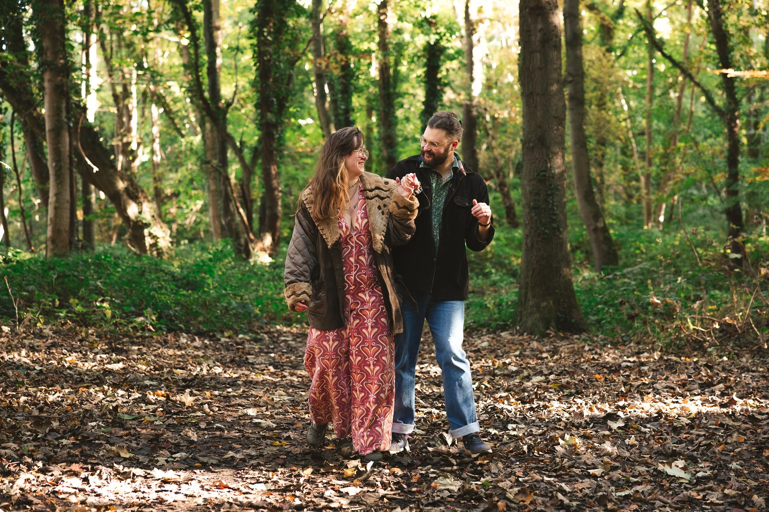 A man and woman walking hand in hand through a forested area, smiling and looking at each other, surrounded by green trees and fallen leaves.