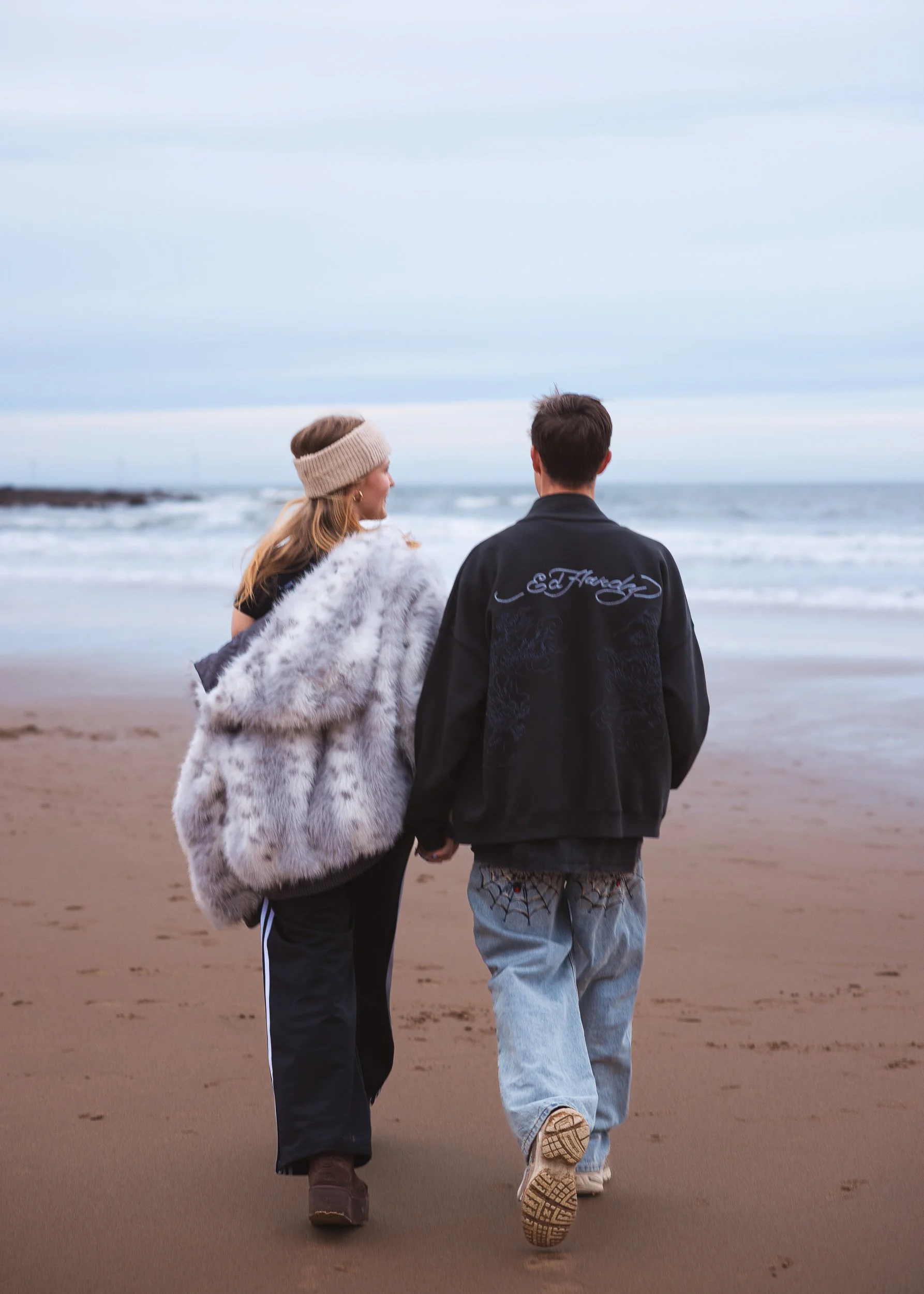 A couple walking hand-in-hand along the beach at sunset, with the woman wearing a fur coat and headband, and the man in a black jacket and jeans, holding hands.
