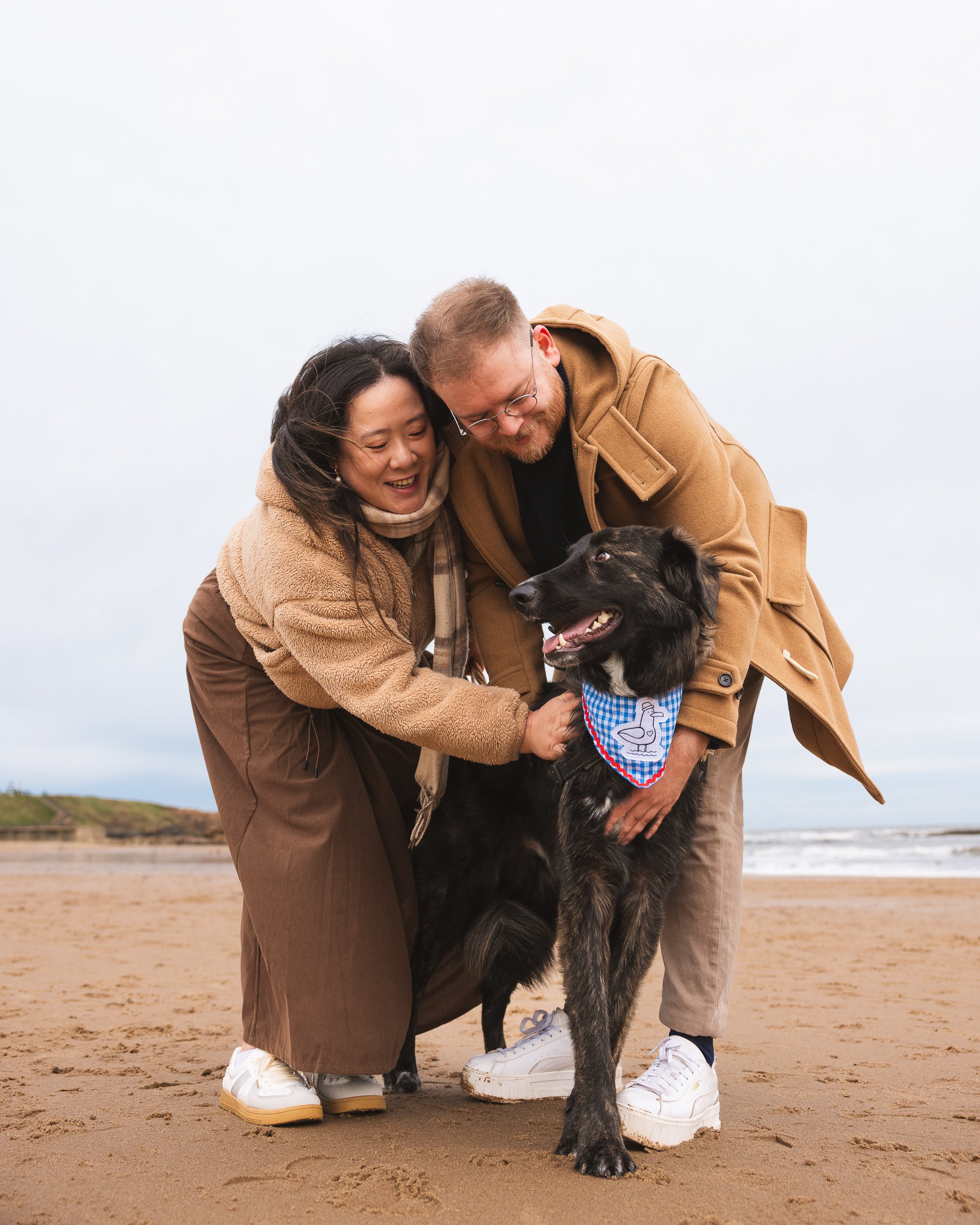 Two people and a dog on a sandy beach. The man and woman are hugging the black dog, which is wearing a blue checkered bandana.