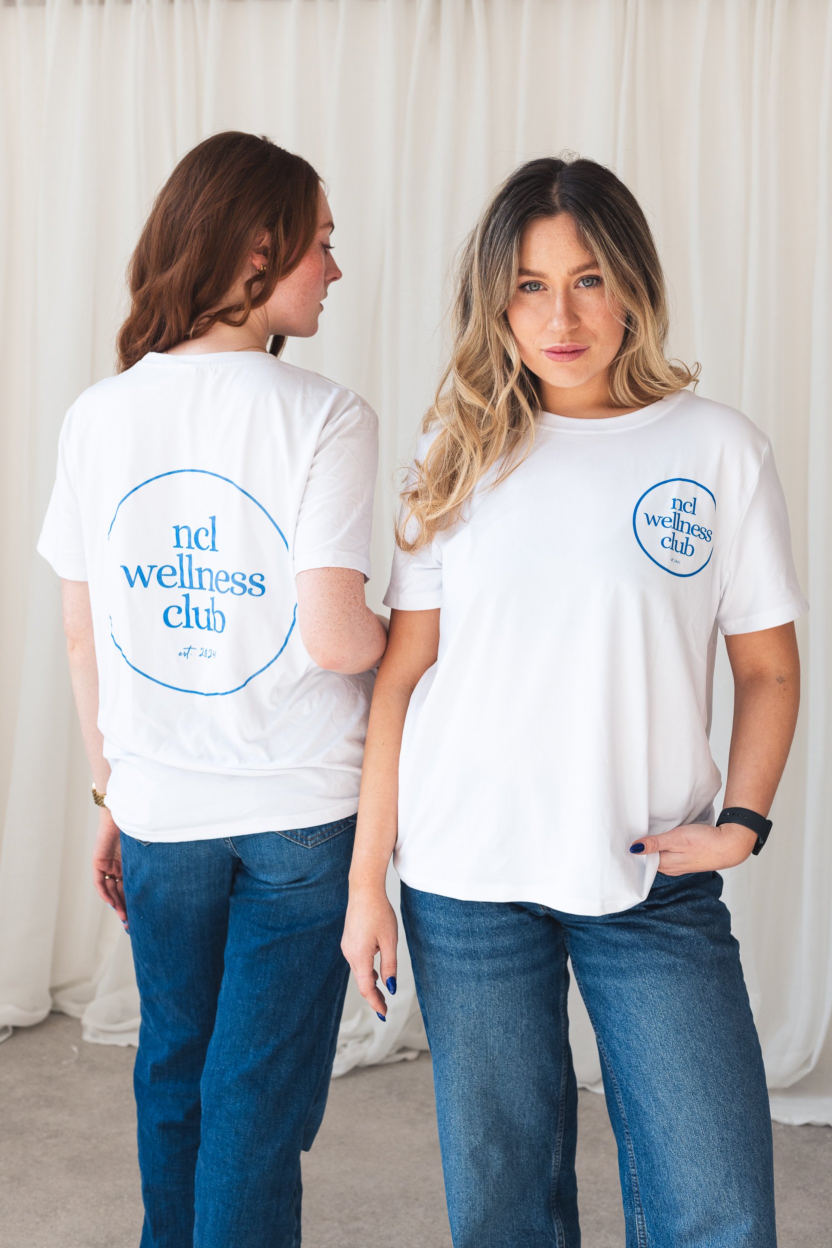 Two women wearing white T-shirts with blue 'ncl wellness club' logos posing against a cream-colored curtain backdrop. One woman faces forward, while the other has her back turned.