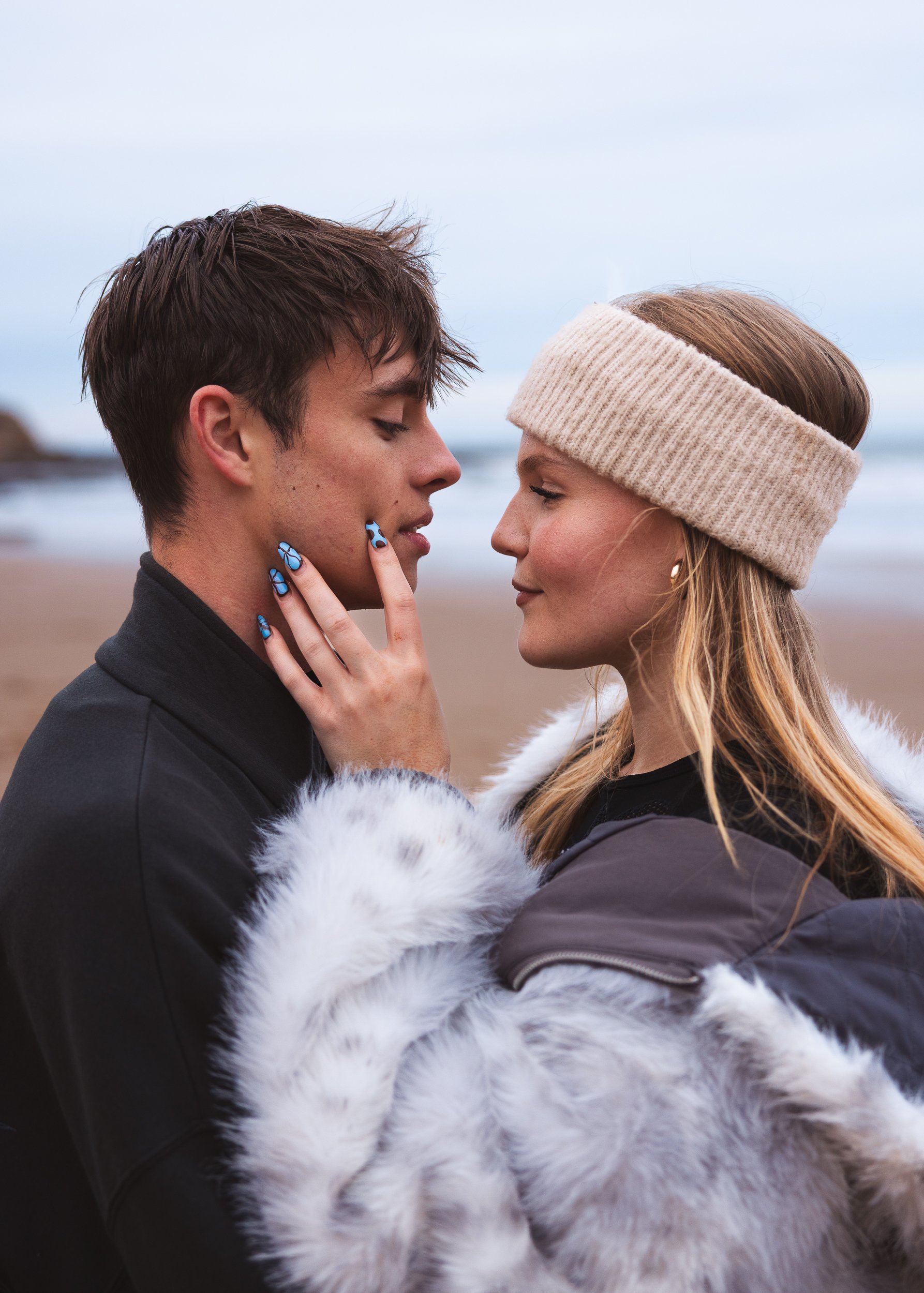 A young man and woman are standing close to each other on the beach; they are touching foreheads and eyes closed, with gentle expressions. The woman wears a beige headband, and the man has short, dark hair. She is dressed in a dark coat with white fu