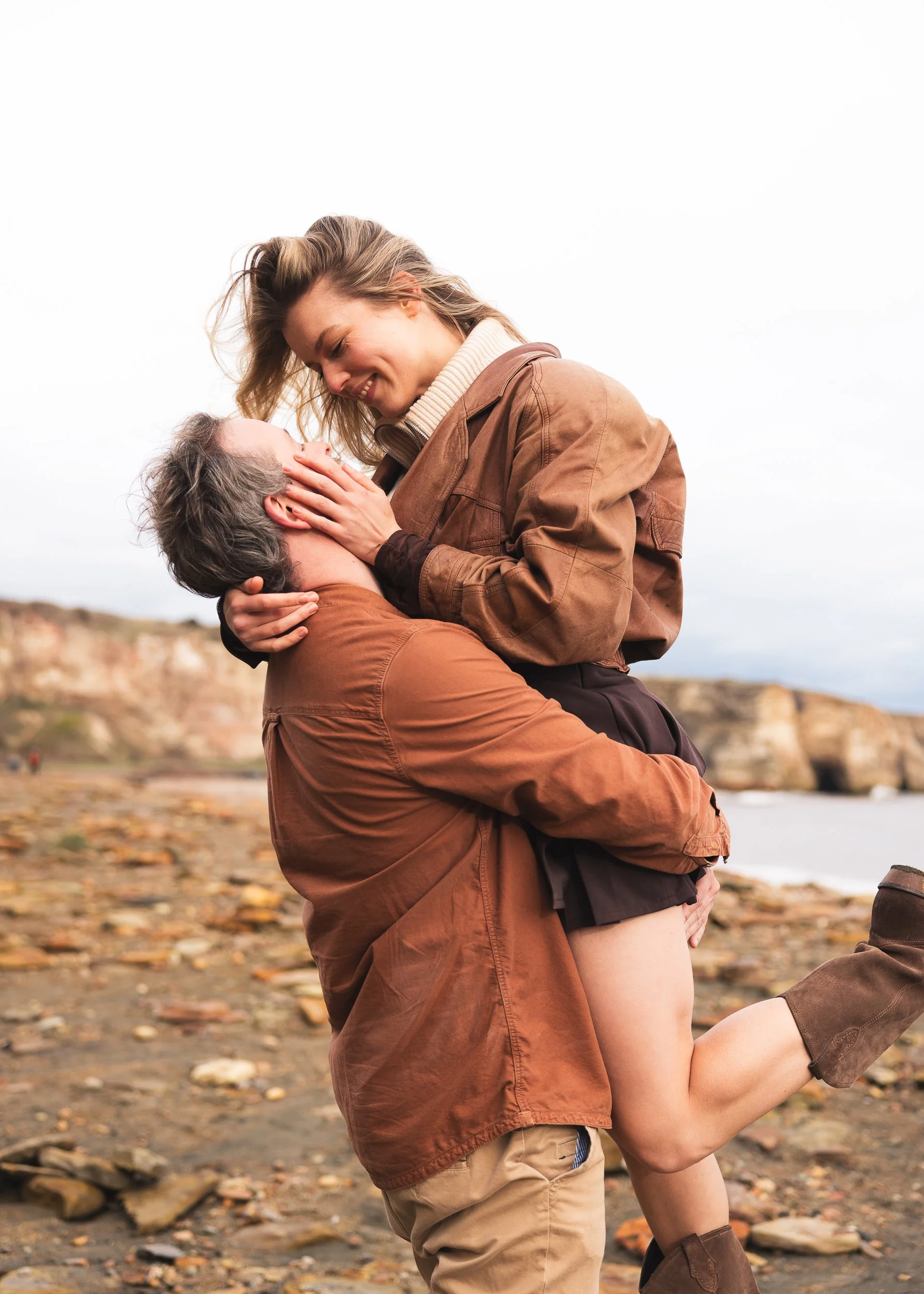 A young woman smiling and holding a man at the beach during autumn, with rocks and fallen leaves on the ground.