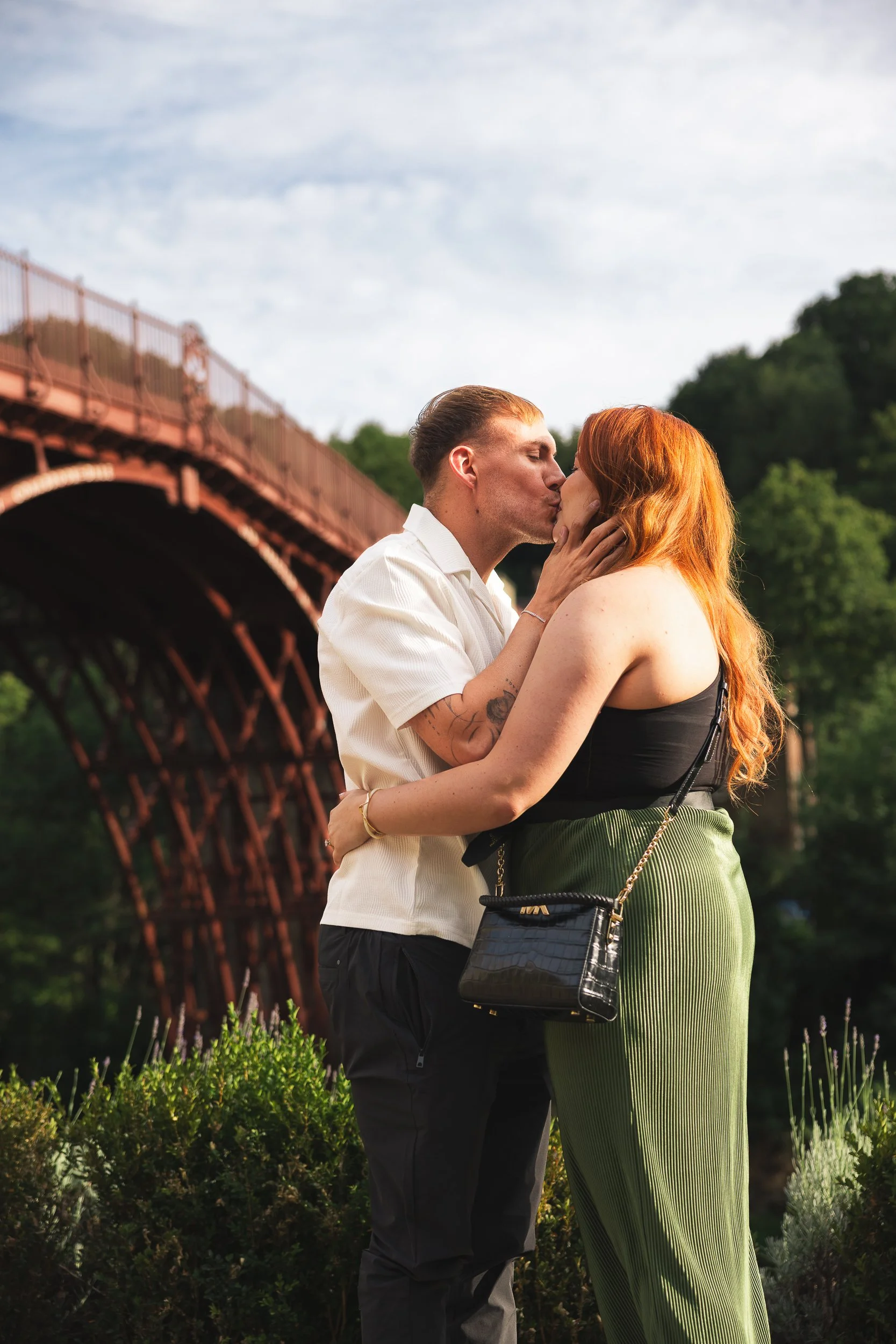 A couple kisses outdoors on a bridge with greenery in the background.