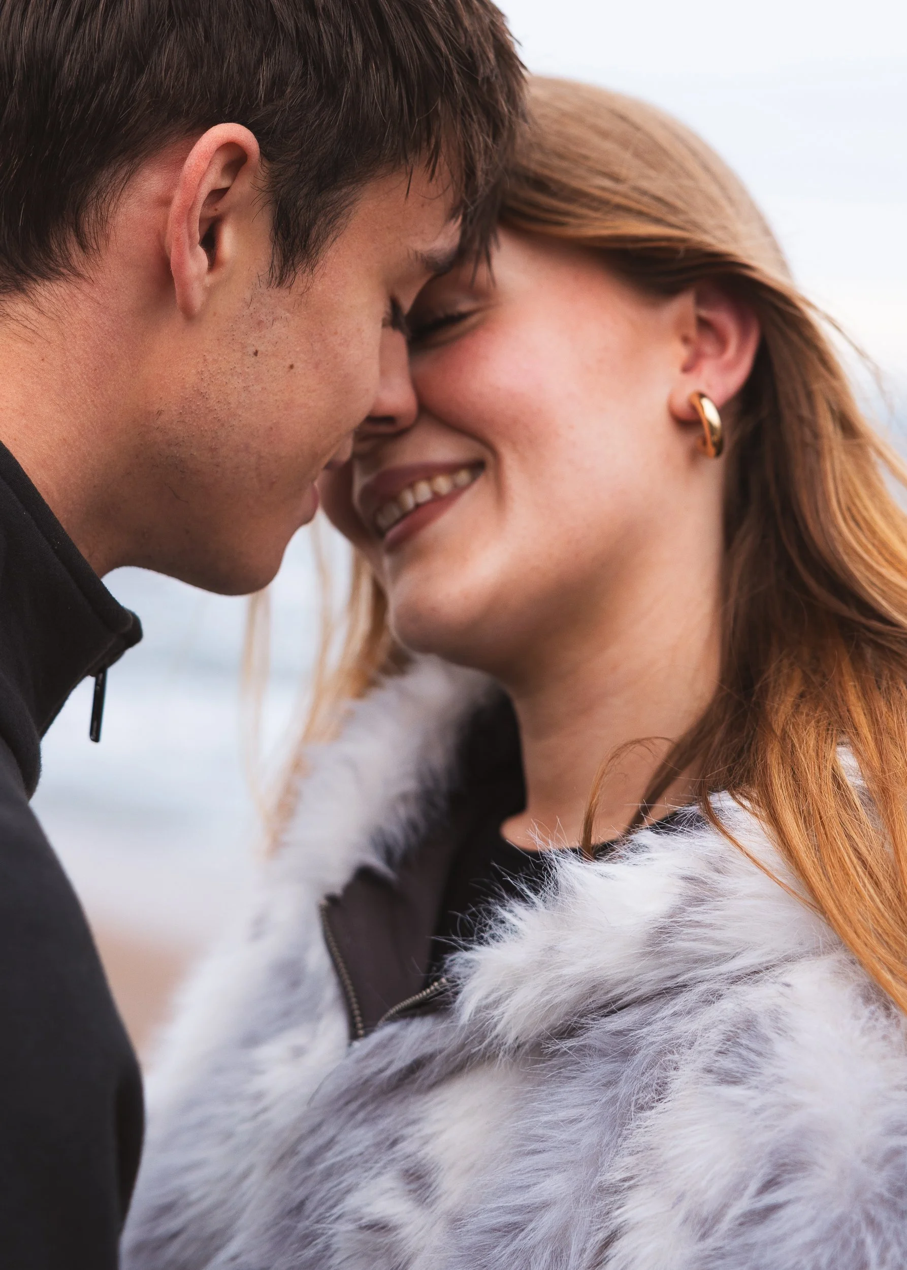 A man and a woman close together with foreheads touching, smiling and enjoying a moment outdoors. The woman has red hair and is wearing a fur-lined jacket and a gold earring. The man has short brown hair and is wearing a black jacket.