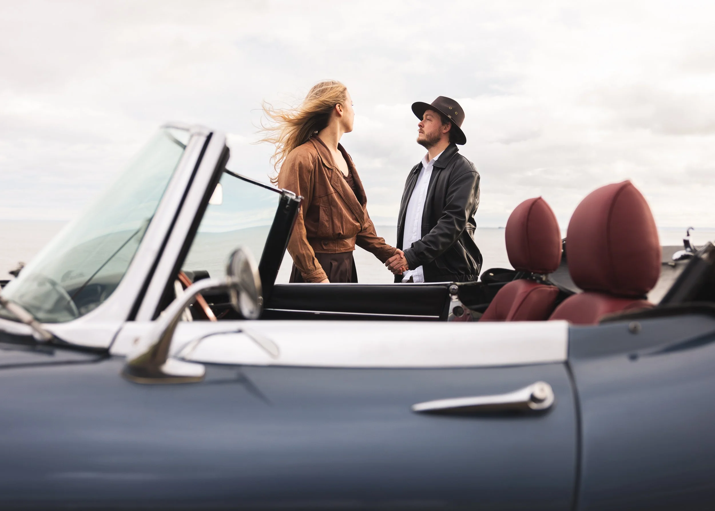 A man and woman holding hands and looking at each other, standing outside near a convertible car on a cloudy day.