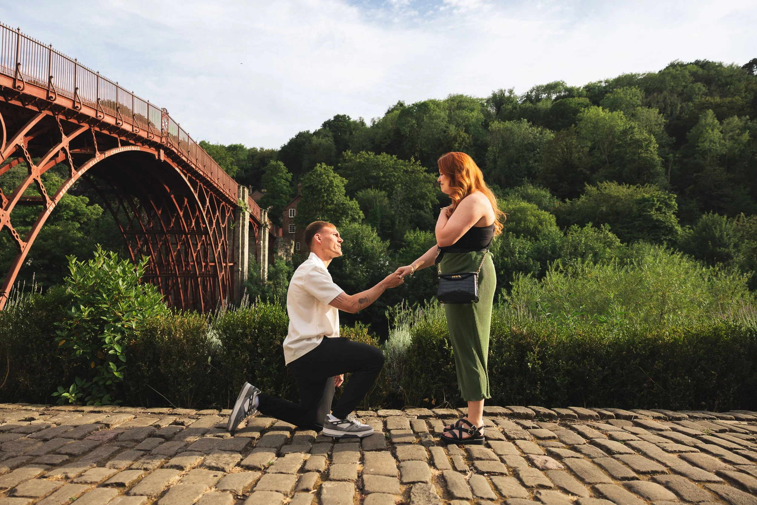 A man kneeling on one knee on a cobblestone path, holding hands with a woman standing in front of him; she is smiling and looking at him, with a lush green hill and an iron bridge in the background.