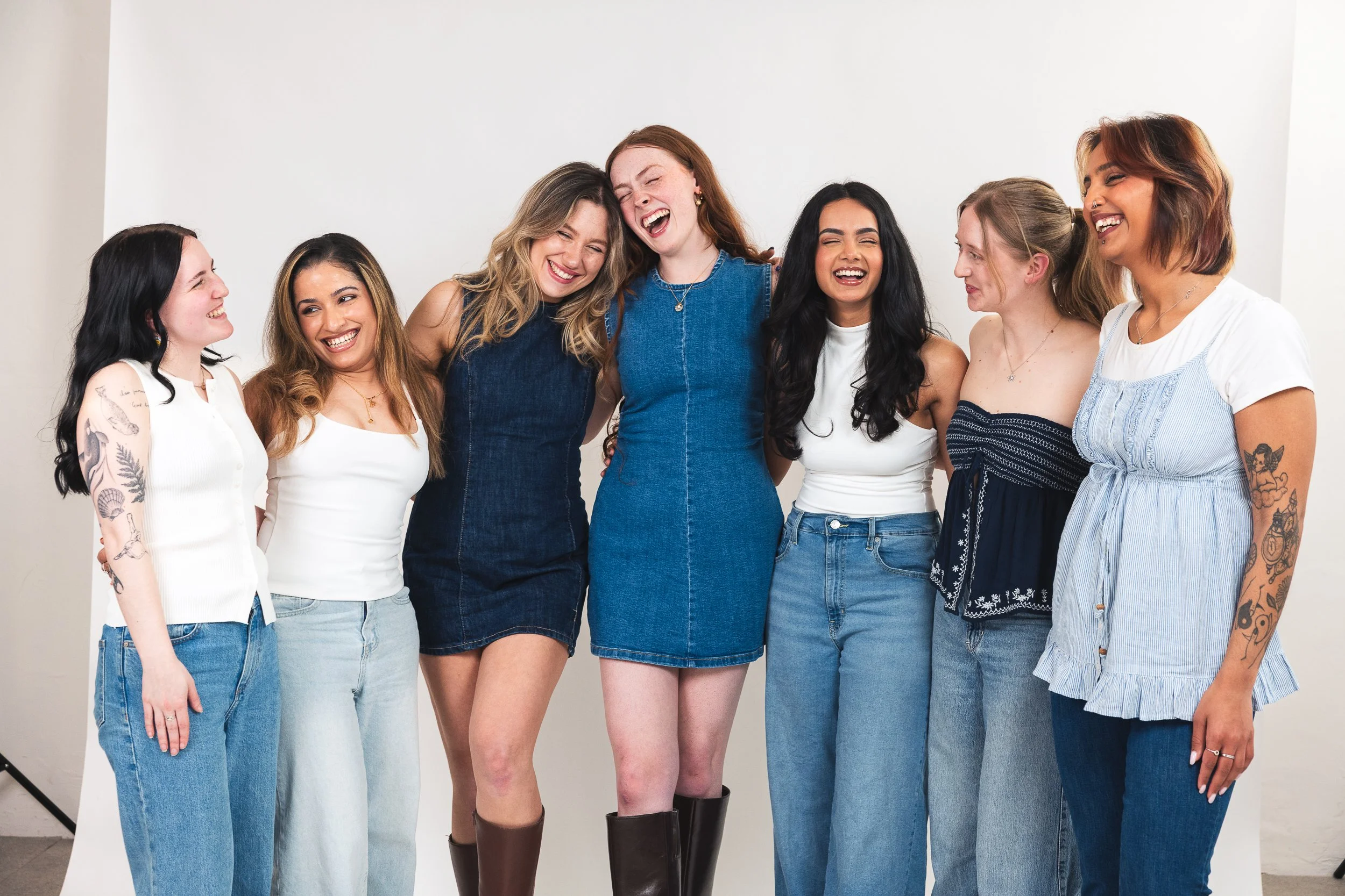 Group of seven young women standing together, smiling and laughing, posing against a plain white background. They are wearing casual summer clothes and have various tattoos and hairstyles.