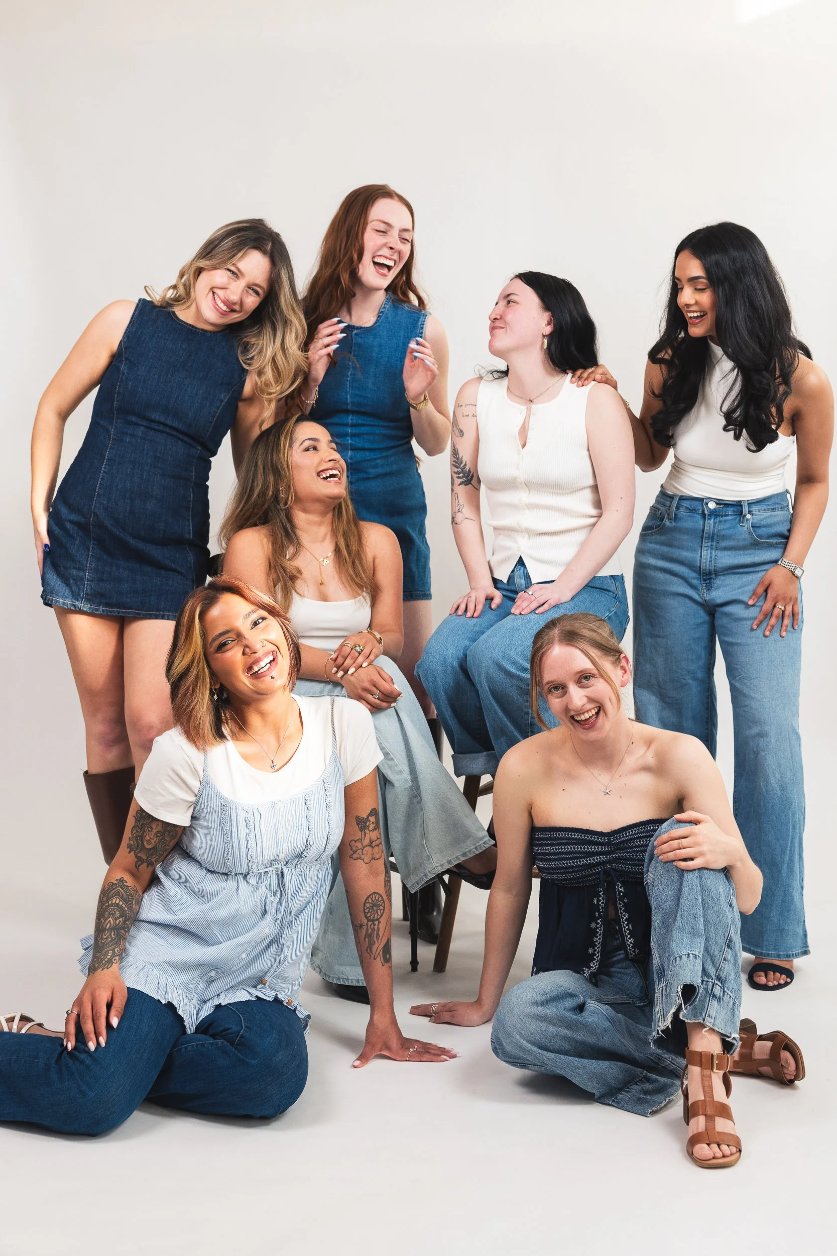 Group of seven women with diverse appearances and styles, smiling and laughing together in a studio with plain white background. Some are seated while others stand or crouch, showing camaraderie and joy.