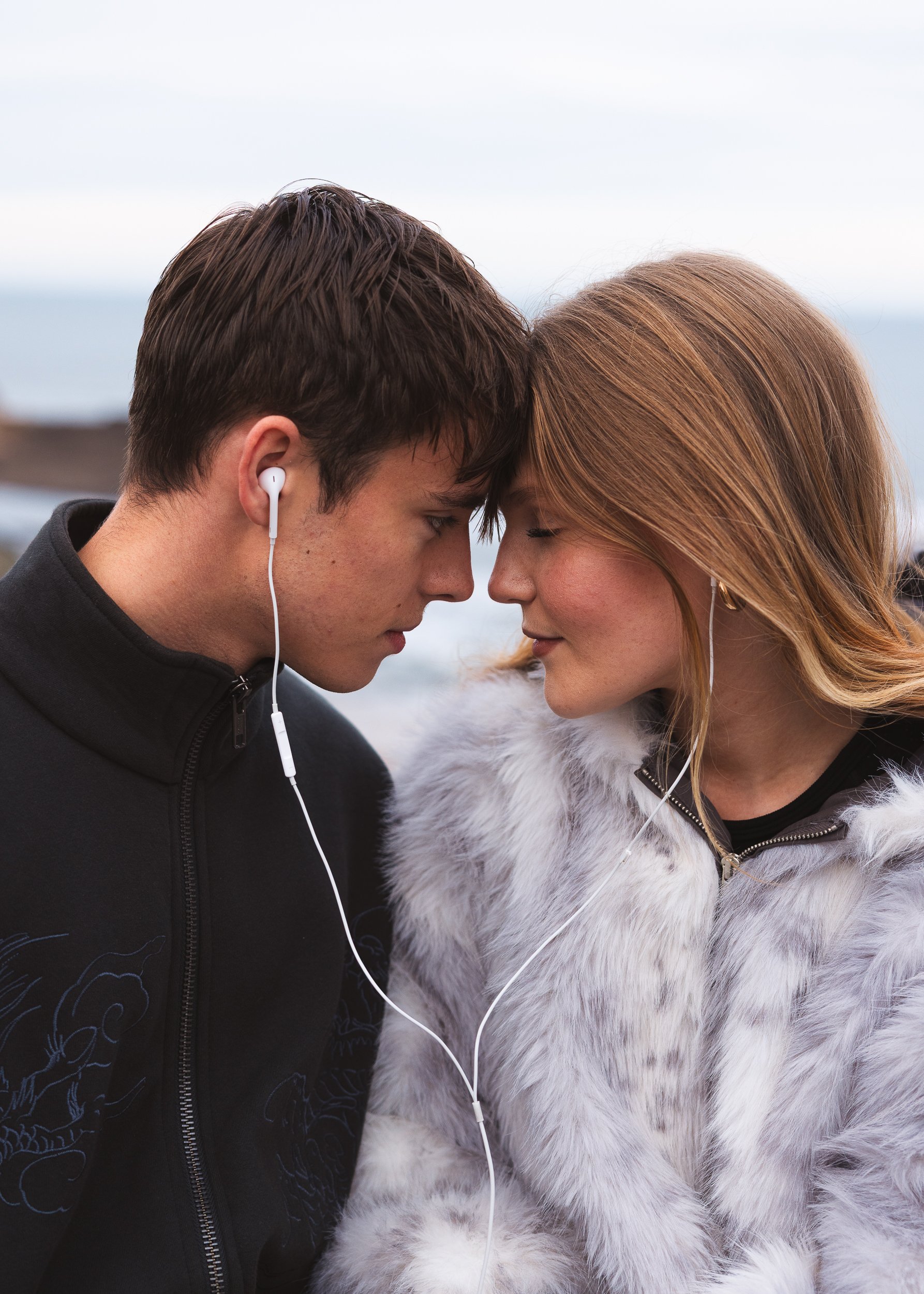 A young man and woman with their foreheads touching, eyes closed, listening to music through earbuds.