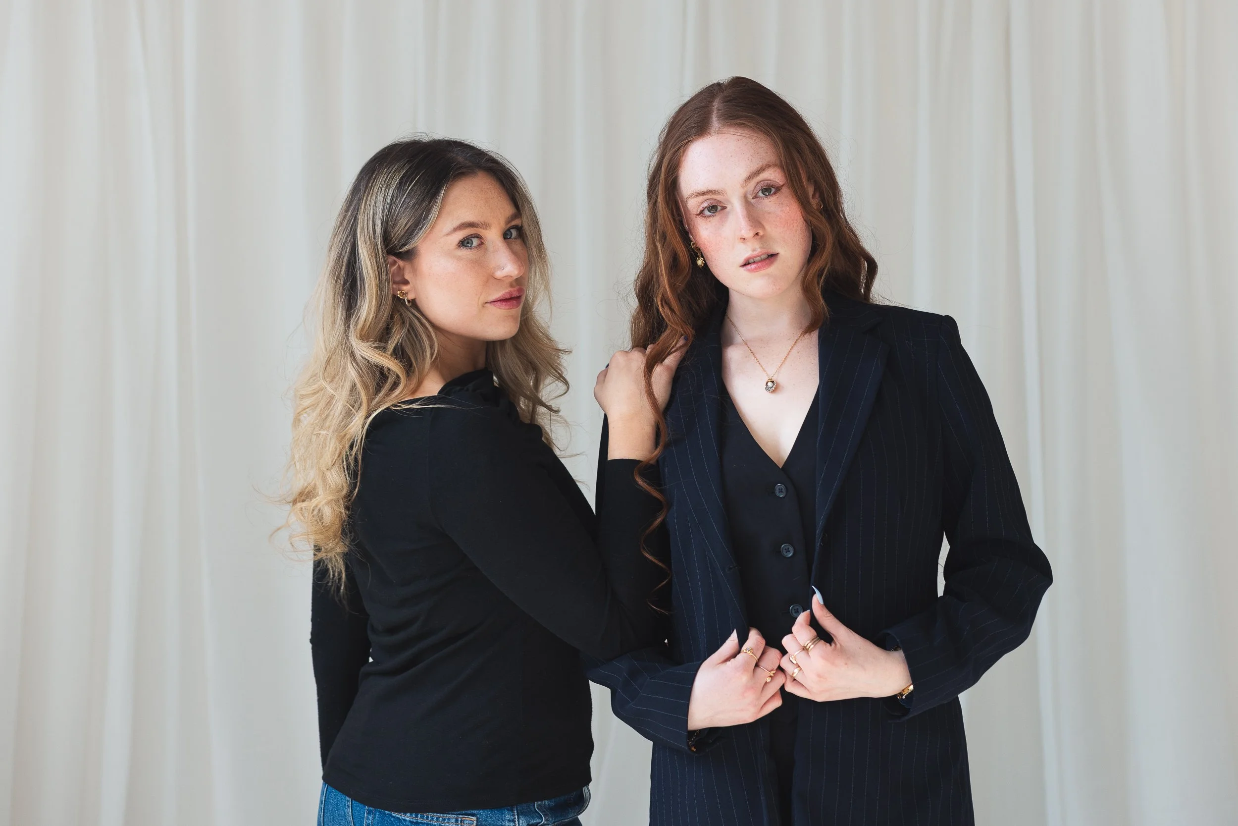 Two young women with light skin standing close together in front of a white curtain, one with long blonde hair and the other with long red hair, both dressed in dark clothing. The red-haired woman is touching her hair and looking at the camera, while