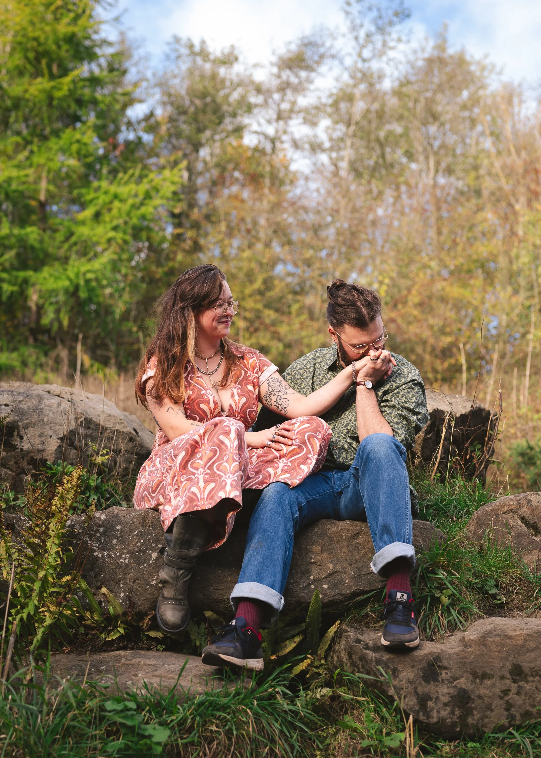 A woman and a man sitting on rocks outdoors, the woman is smiling and appears to be touching the man's hand, which is near her face. The background shows trees with green and some brown leaves, indicating fall. Both are dressed casually, wearing glasses, and seem to be enjoying a moment together in nature.