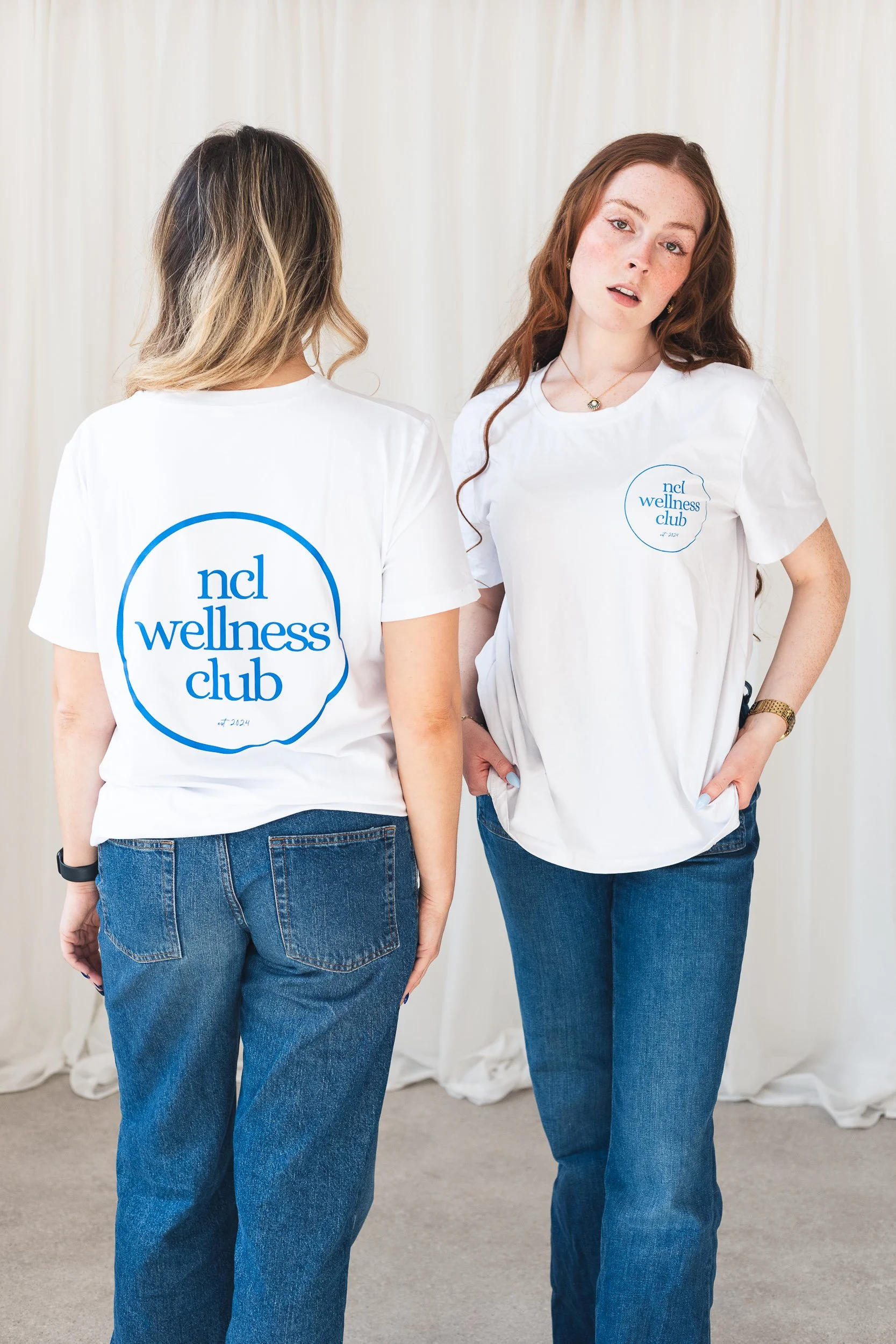 Two women standing side by side wearing white t-shirts with the blue text 'ncl wellness club' printed on the front, facing different directions against a light-colored background.