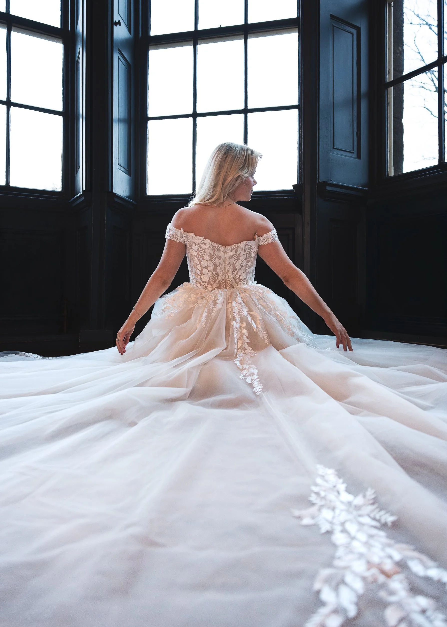A woman in a wedding dress sitting on the floor next to large windows, with a trail of white flower petals behind her on the white floor.