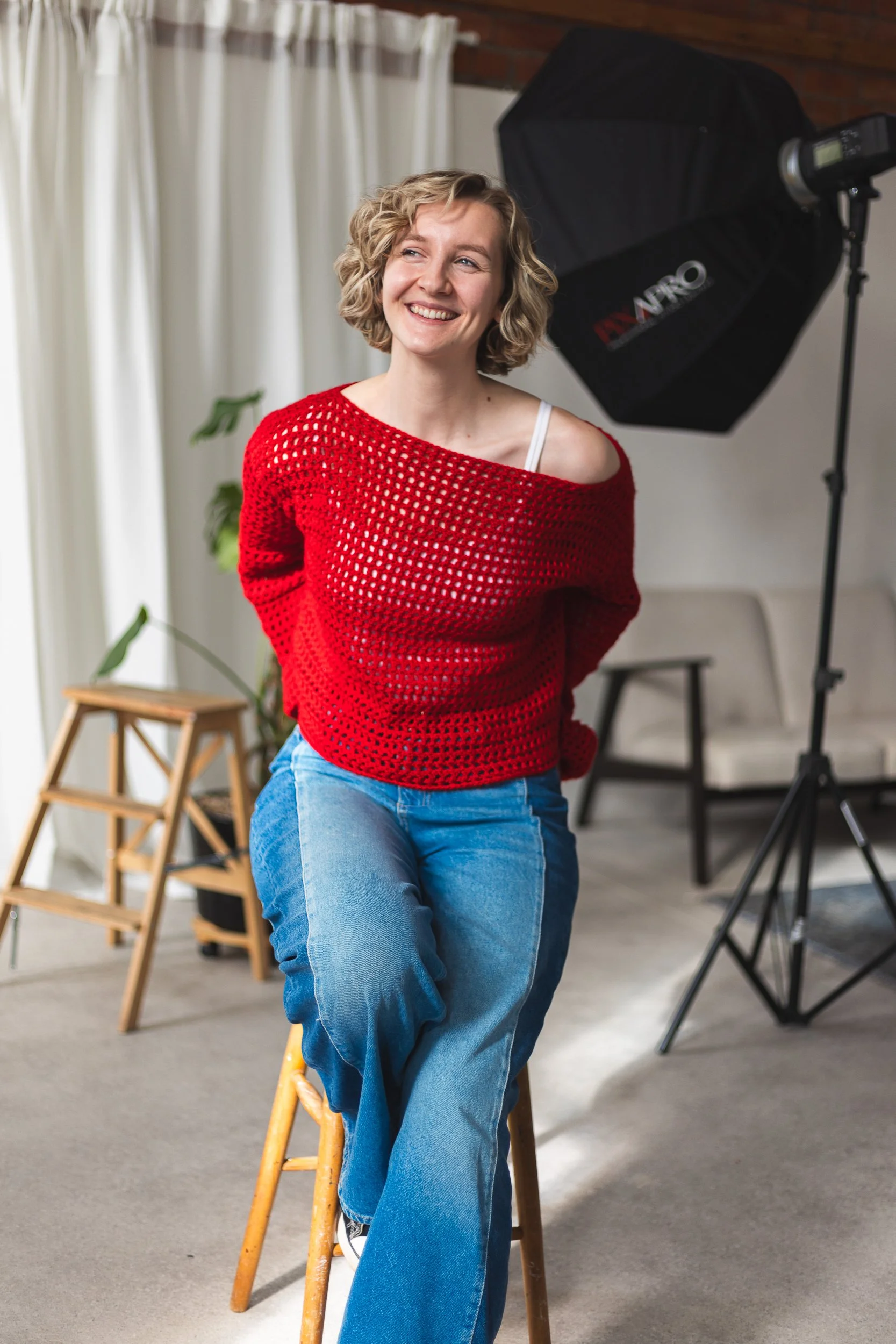 A woman with curly blonde hair, wearing a red open-knit sweater, white tank top, and blue jeans, smiling while seated on a yellow wooden stool in a photography studio with professional lighting equipment.