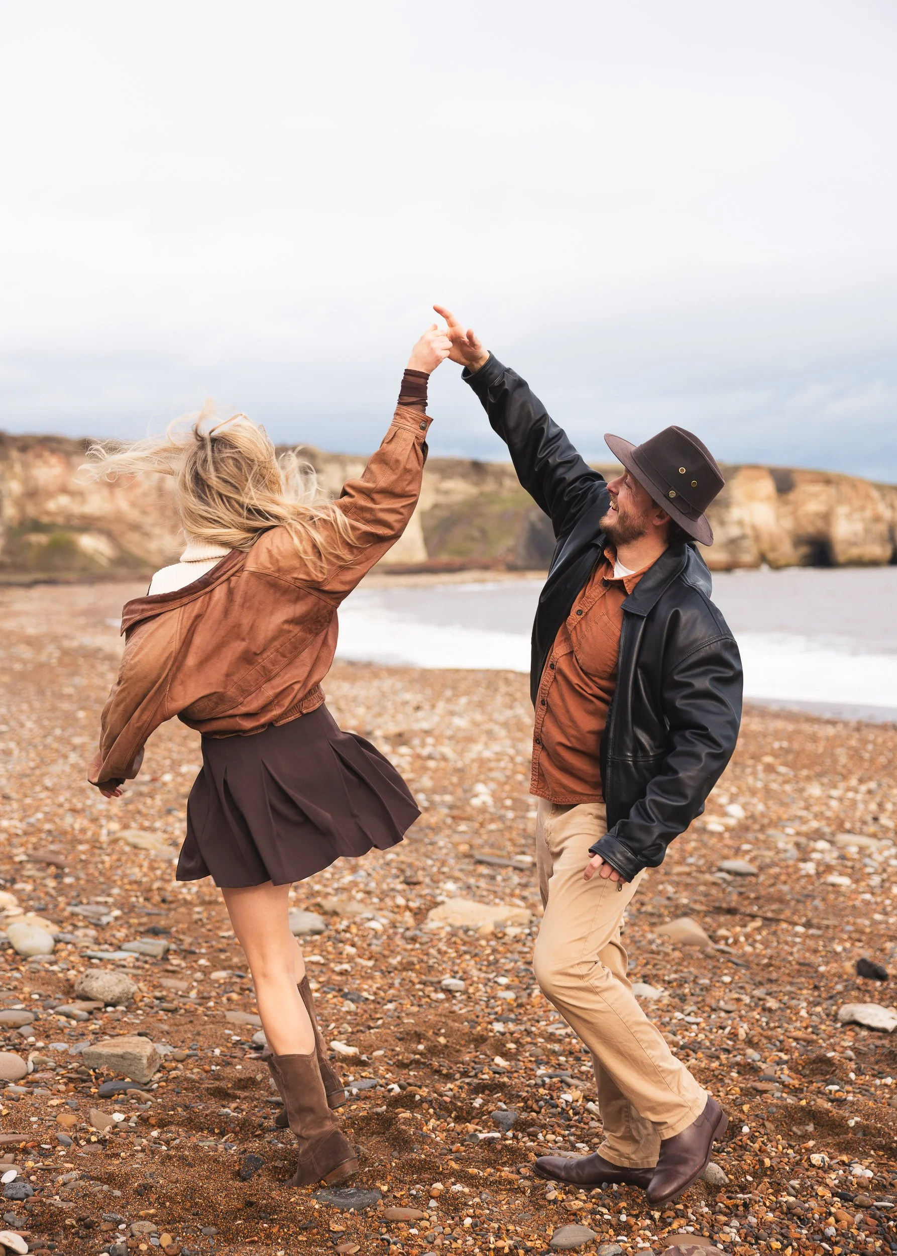 A couple dancing and holding hands on a rocky beach with cliffs in the background, both smiling and enjoying the moment.