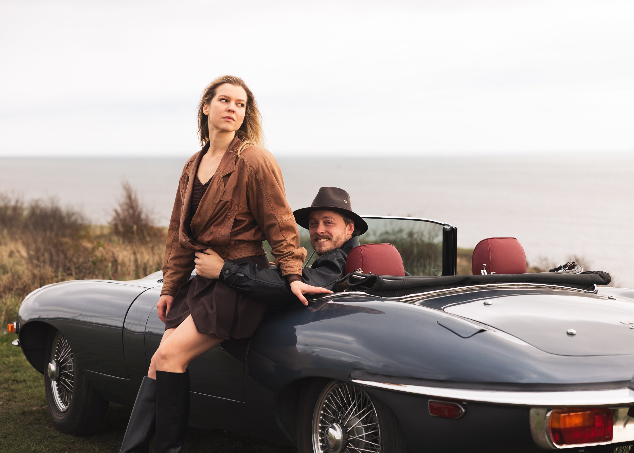 A woman in a brown jacket and black boots leaning against a classic black convertible car with red seats. A man in a black leather jacket and hat is sitting in the driver’s seat smiling. They are outdoors near water with cloudy skies.