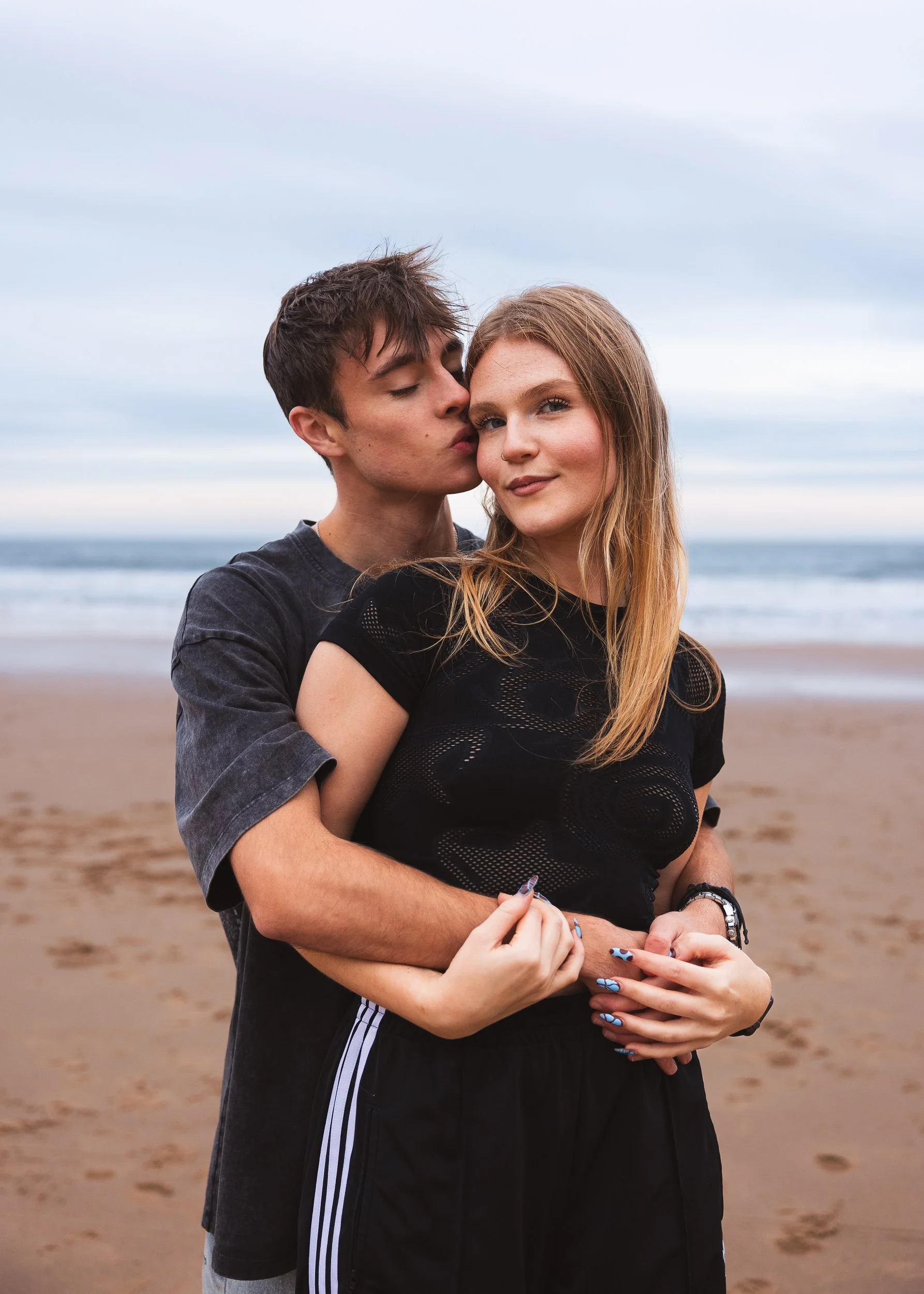 A young man kisses a young woman on the cheek while standing on the beach. The woman is smiling and has long blonde hair, while the man has short brown hair. They are embracing each other with arms around each other. The ocean and cloudy sky are in t
