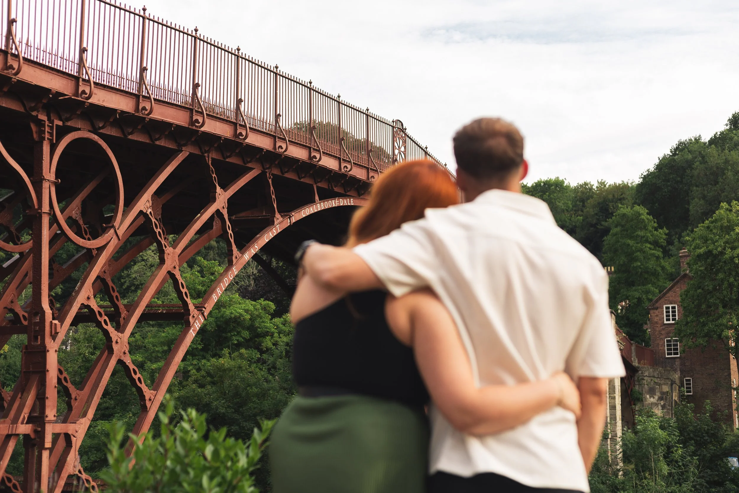 A couple embracing outdoors in front of a red iron bridge with greenery and houses in the background.