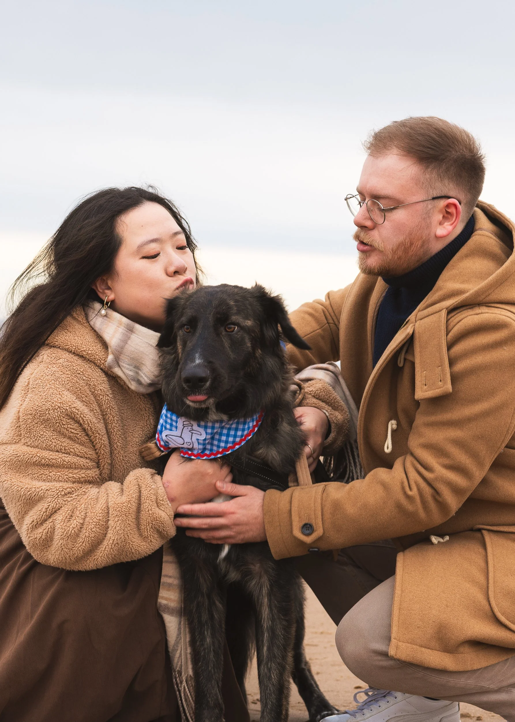 Two people, a woman and a man, are outdoors holding a black dog with a blue and white bandana. The woman has long dark hair and is wearing a brown cozy jacket. The man has short hair, glasses, and is wearing a tan coat. They are close to each other, appearing to share a moment with the dog.