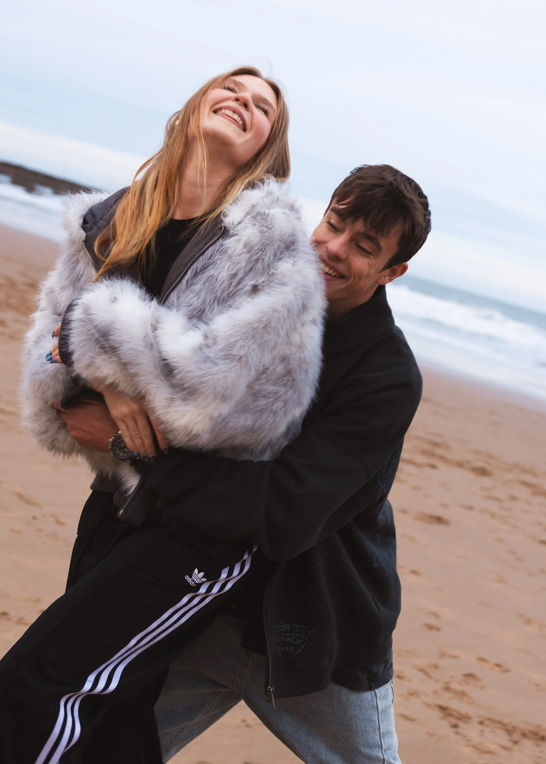 A man and a woman smiling and hugging on a sandy beach with waves in the background.