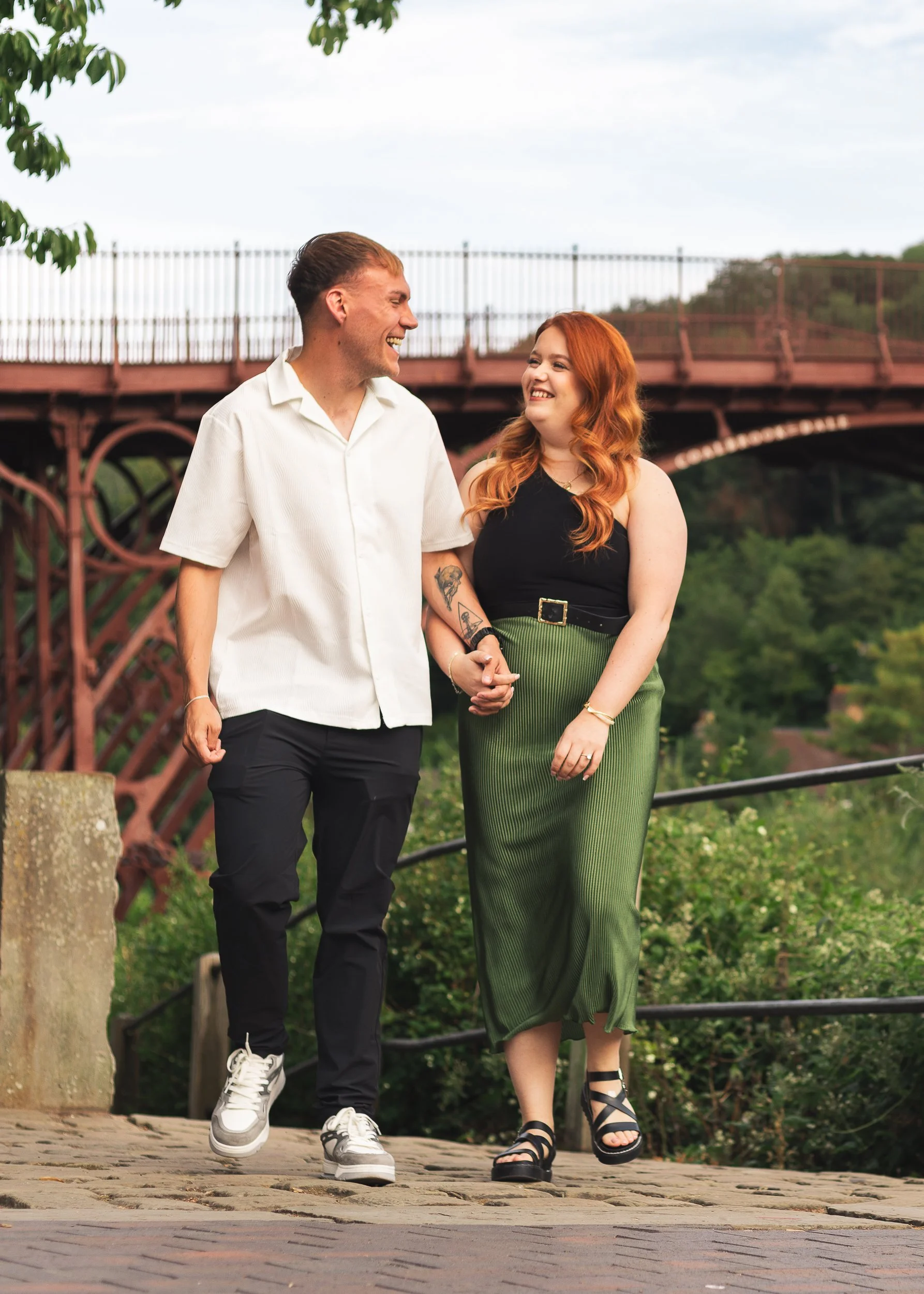 A couple walking hand-in-hand on a cobblestone path, smiling at each other. The man has light skin, short brown hair, and is wearing a white short-sleeved shirt, black pants, and sneakers. The woman has light skin, long red hair, and is wearing a bla