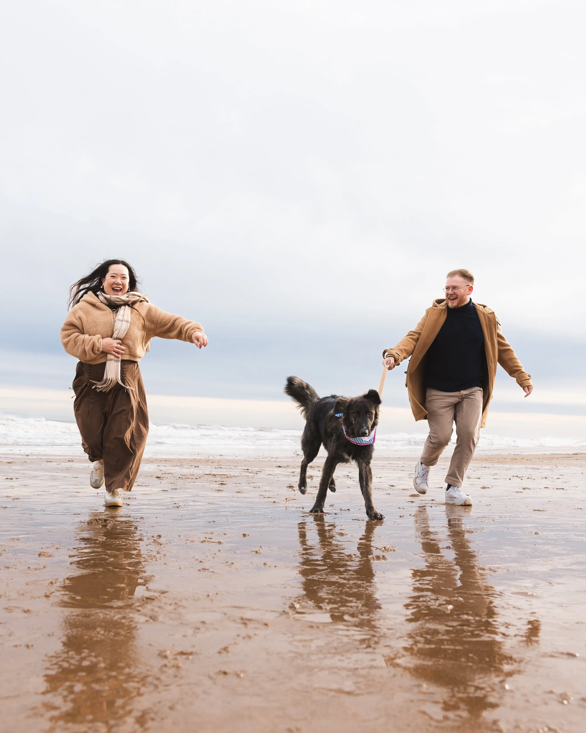 Two people, a woman and a man, playing with a dog on a beach. The woman is wearing a brown jacket and pants, the man is wearing a brown coat and beige pants. The dog is black and wearing a bandana. The sky is overcast.