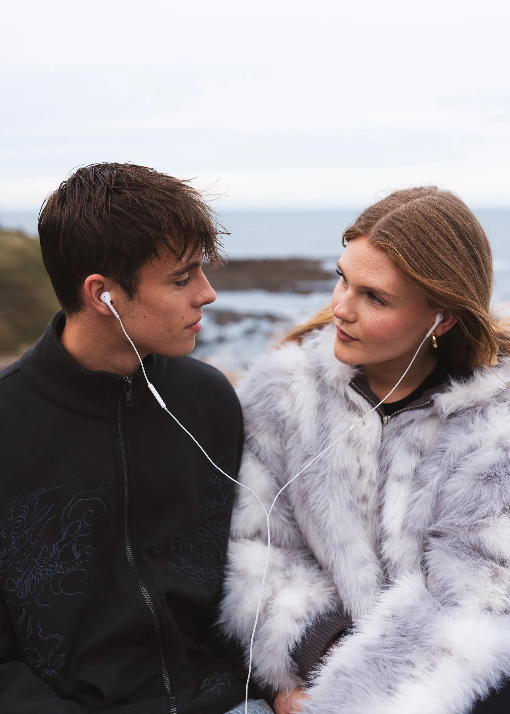 A young man and woman face each other outdoors near the ocean, wearing earbuds and a fur coat, with overcast sky and rocky shoreline in the background.