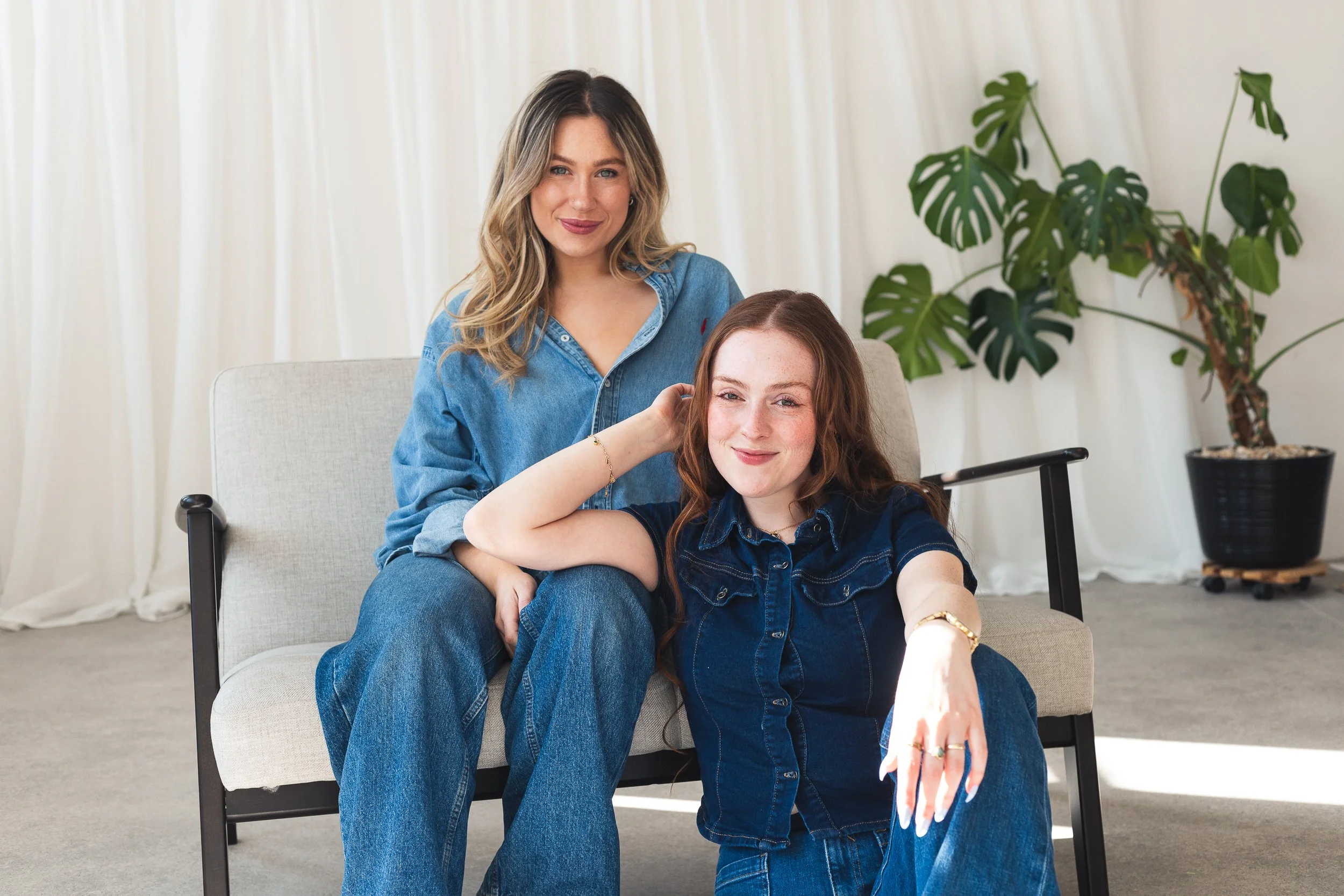 Two women with curly hair posing indoors, one seated on a beige armchair and the other standing behind her. The woman seated has reddish-brown hair and is wearing a denim shirt, while the standing woman has wavy blonde hair and is wearing a denim jacket. They are smiling and appear to be close to each other. There is a large potted plant with broad leaves in the background.