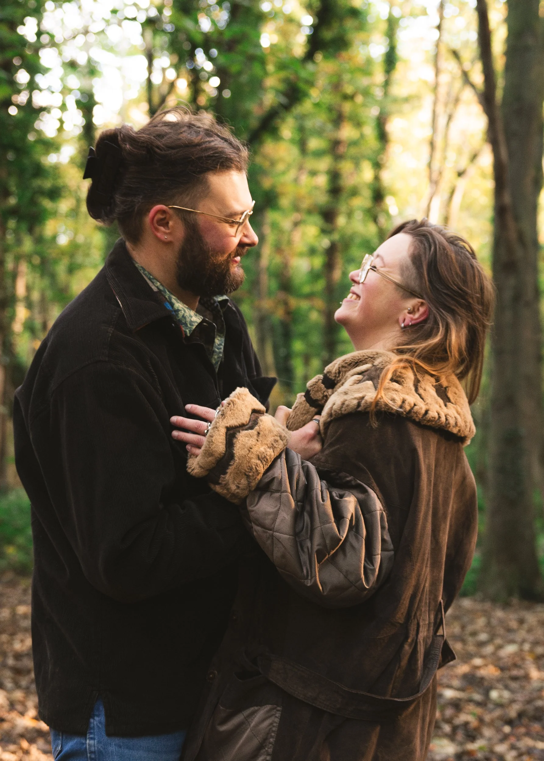 A couple smiling and holding hands in a forest, looking at each other, during autumn.