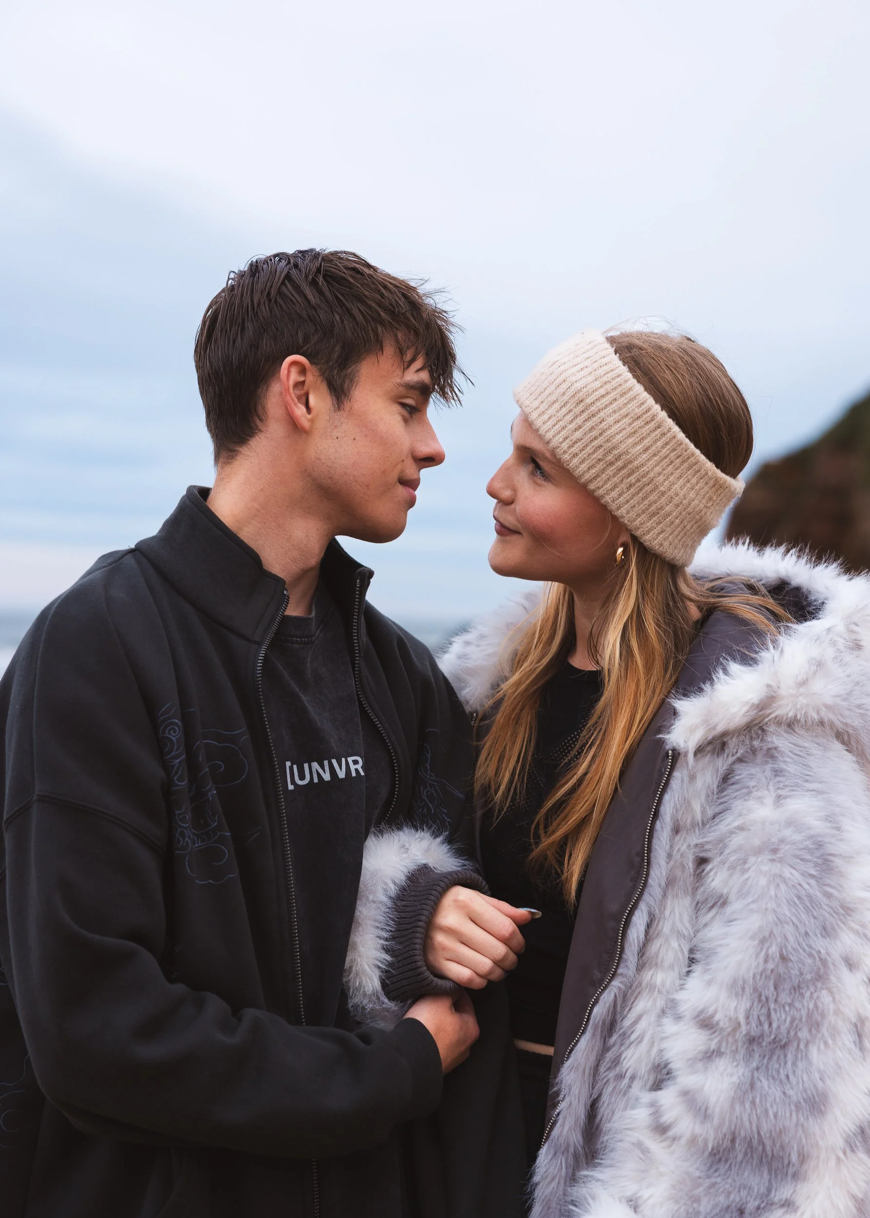 A young man and woman face to face outdoors, holding hands, with the man smiling softly and the woman wearing a beige headband and a fluffy jacket.