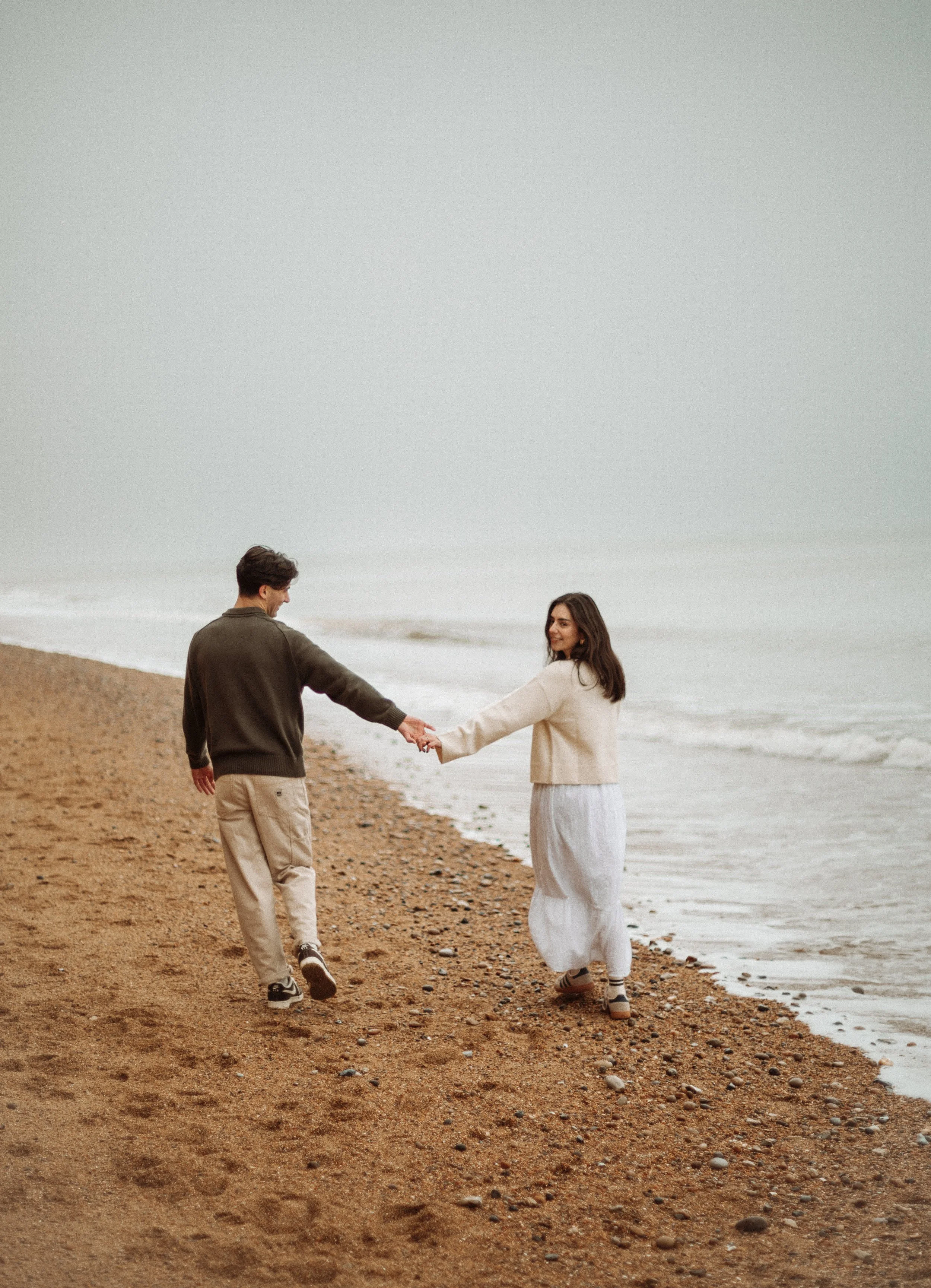 A couple holding hands and walking along a sandy beach near the ocean.