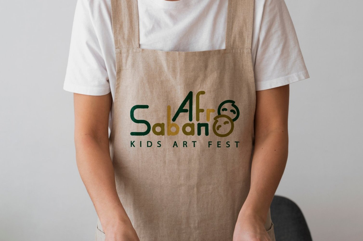 Person wearing a khaki apron with a logo for Kids Art Fest, featuring stylized faces and text reading 'Afro Saban Kids Art Fest' in green and yellow, against a light gray background.