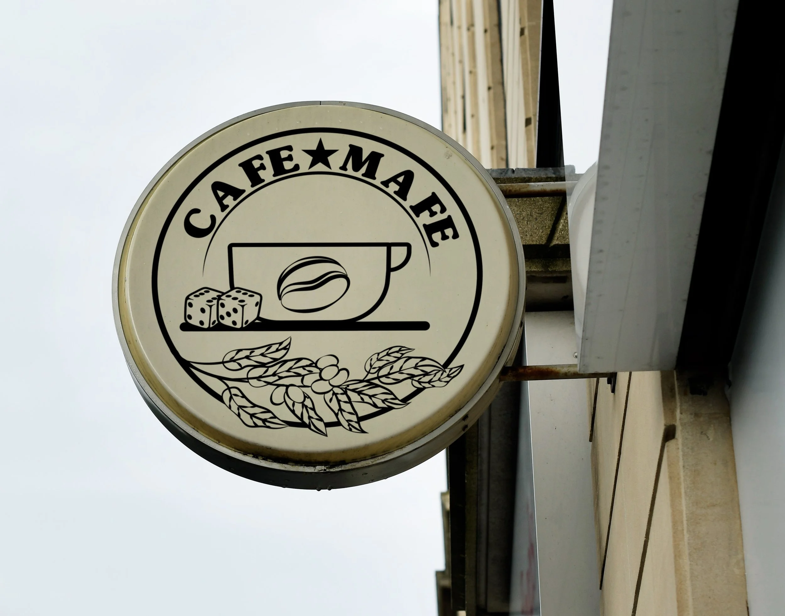Close-up of a circular sign with a black border, featuring the words "Cafe Mafé" and illustrations of dice, a bowl, and wheat, mounted on a building exterior with a wooden structure and cloudy sky in the background.
