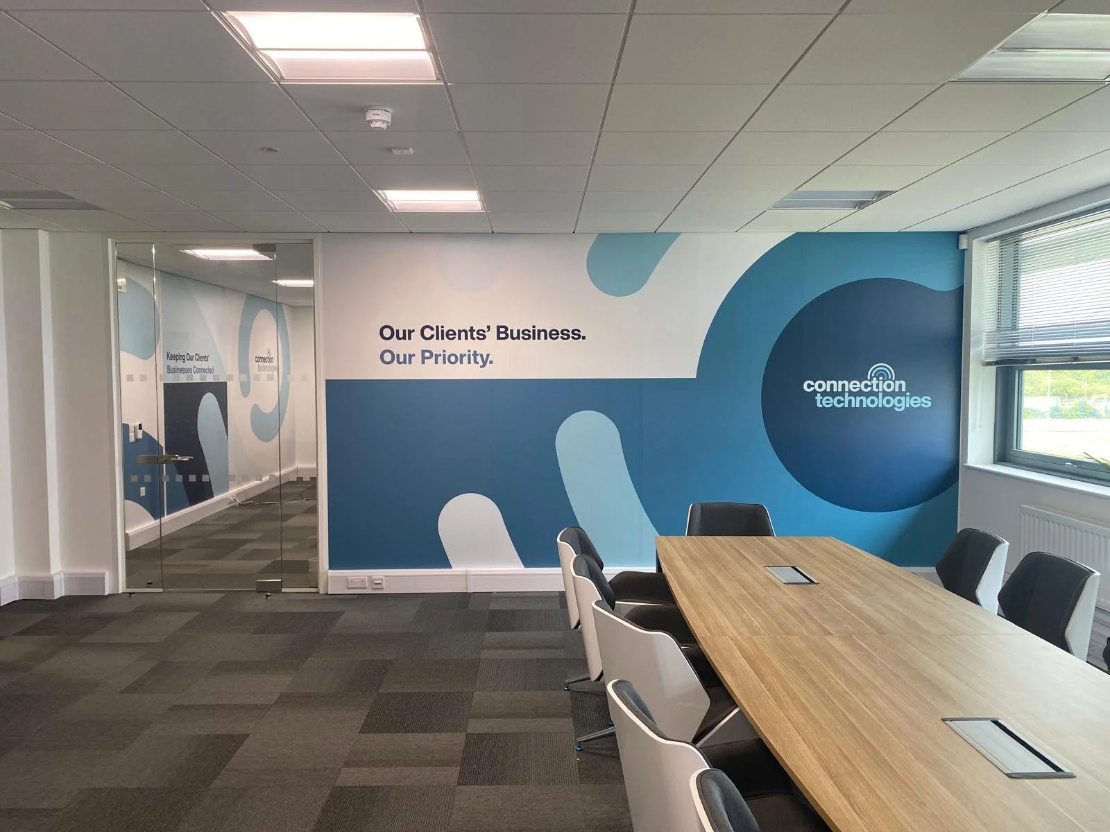 Modern conference room with a wooden table, black and white chairs, large wall mural with blue abstract shapes, and a window with blinds. The mural has the text 'Our Clients' Business. Our Priority.' and 'connection technologies'.