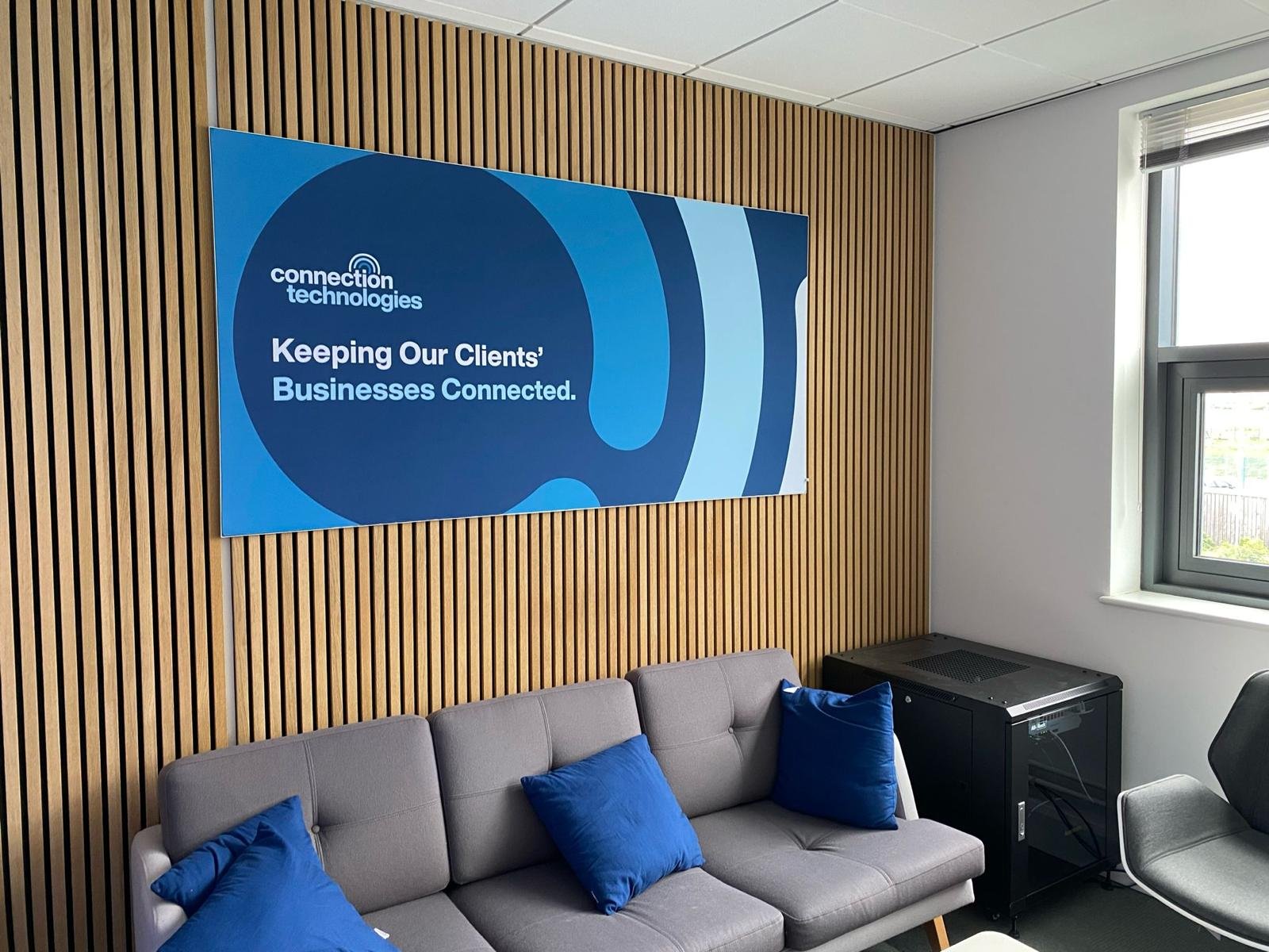 Office lounge area with a grey sofa and blue cushions, a wall-mounted blue sign with logo and text, a window on the right, and a black electronic device underneath the window.