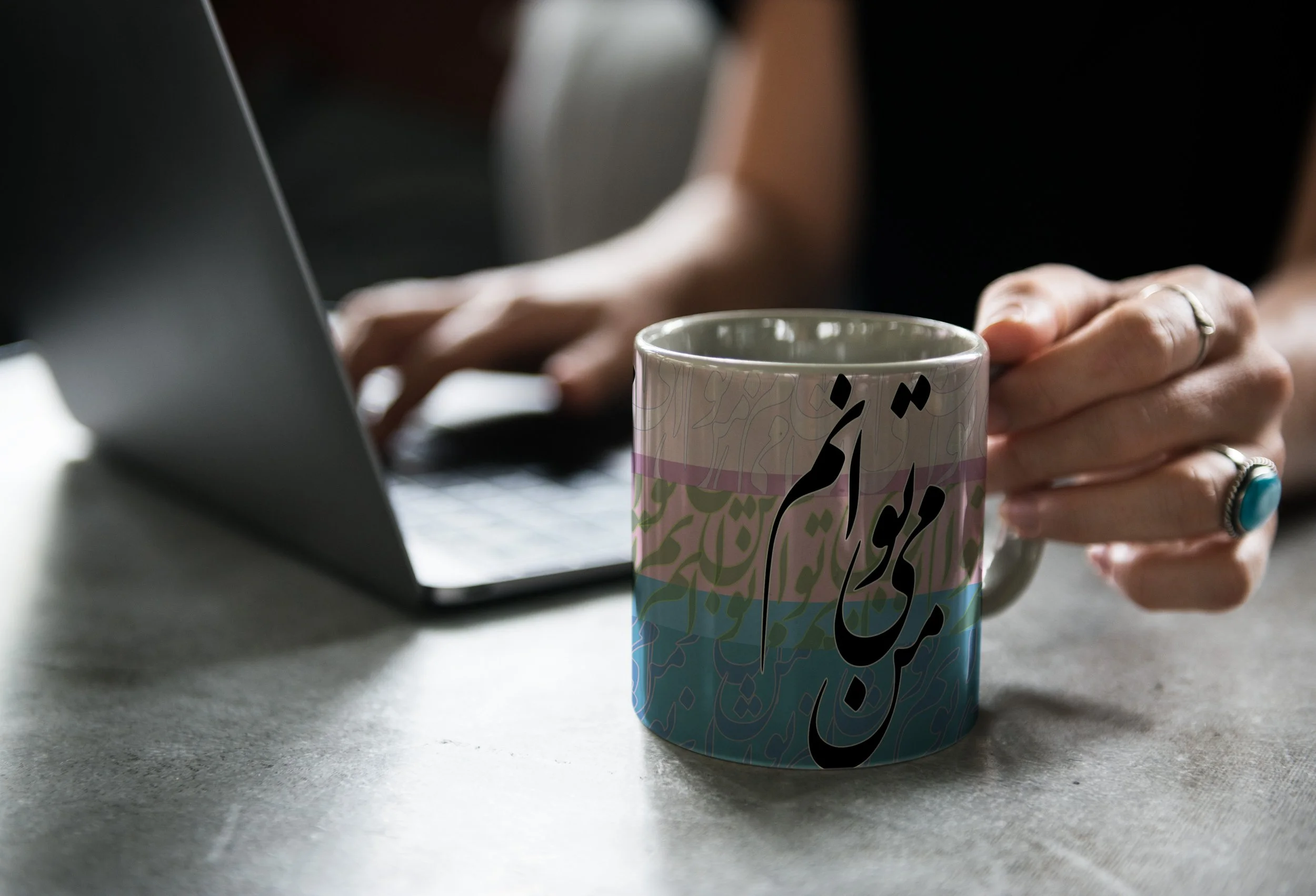 Close-up of a person's hand holding a colorful mug with abstract art, with a laptop in the background on a gray surface.